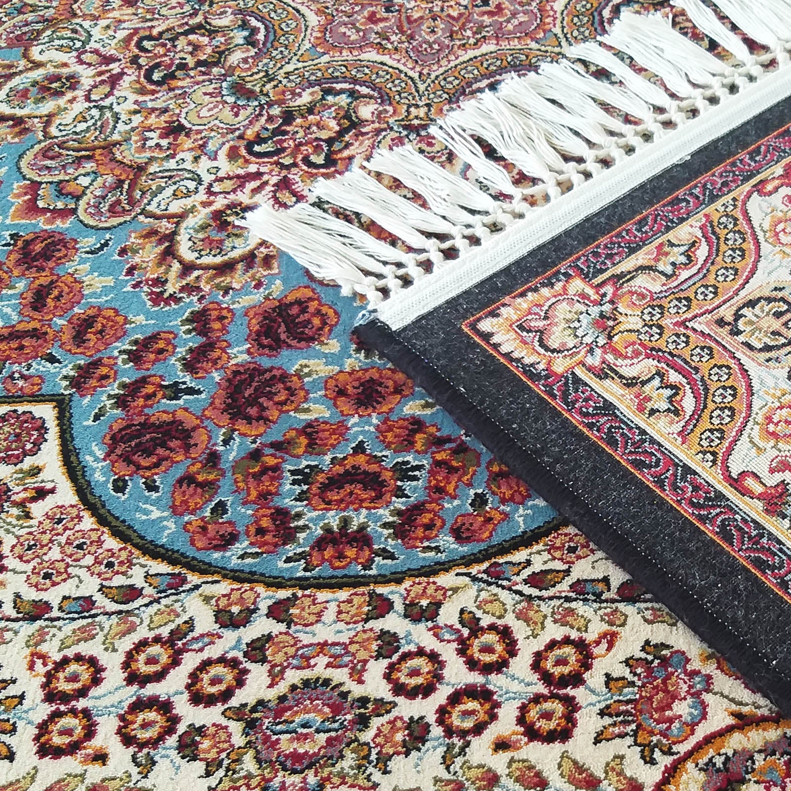Tapis aux motifs floraux et cachemire, richement coloré. Il présente des franges blanches et une partie repliée montrant son envers foncé