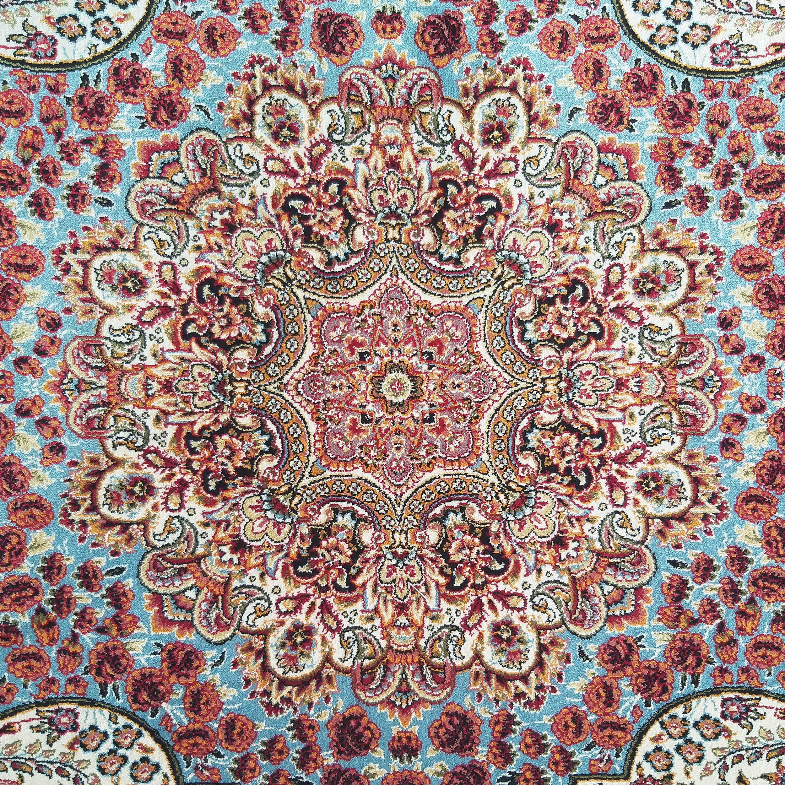 Tapis floral orné. Le tapis affiche un grand médaillon central rouge, crème et orange sur un fond bleu clair parsemé de roses