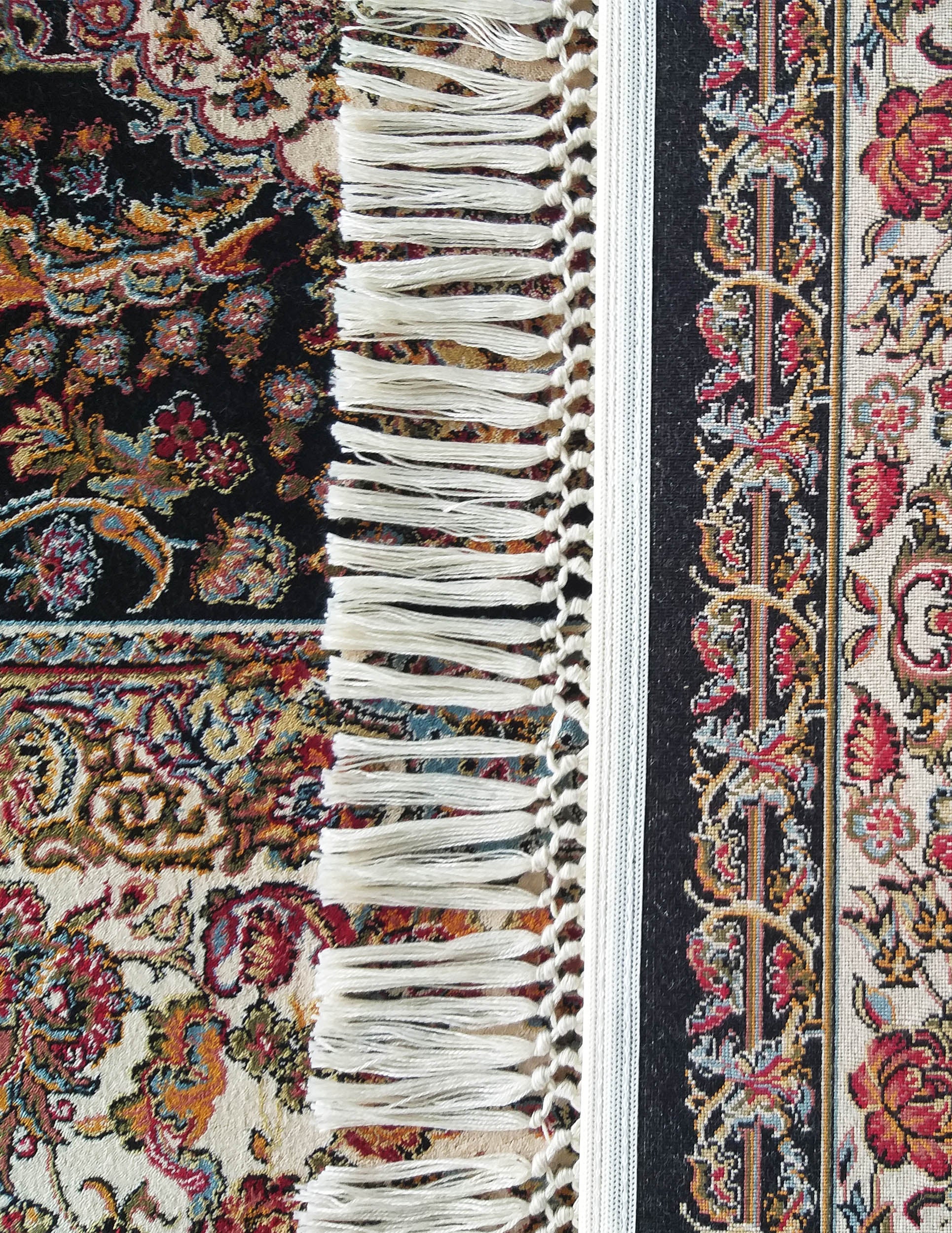 Tapis aux motifs floraux rouges, bleus et oranges. Le fond est noir à gauche et crème à droite, avec une frange blanche centrale. Une étiquette noire est visible
