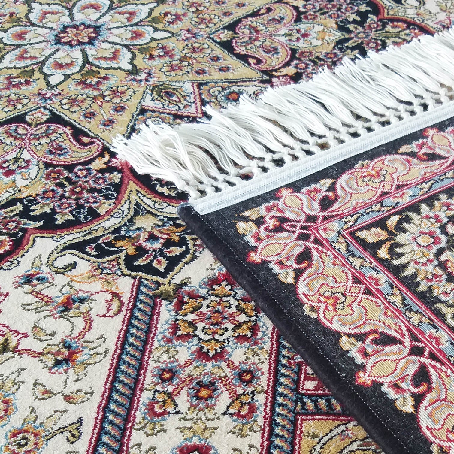 Tapis aux riches motifs floraux et couleurs crème, noir, doré, rouge, bleu, des franges blanches visibles sur le bord enroulé