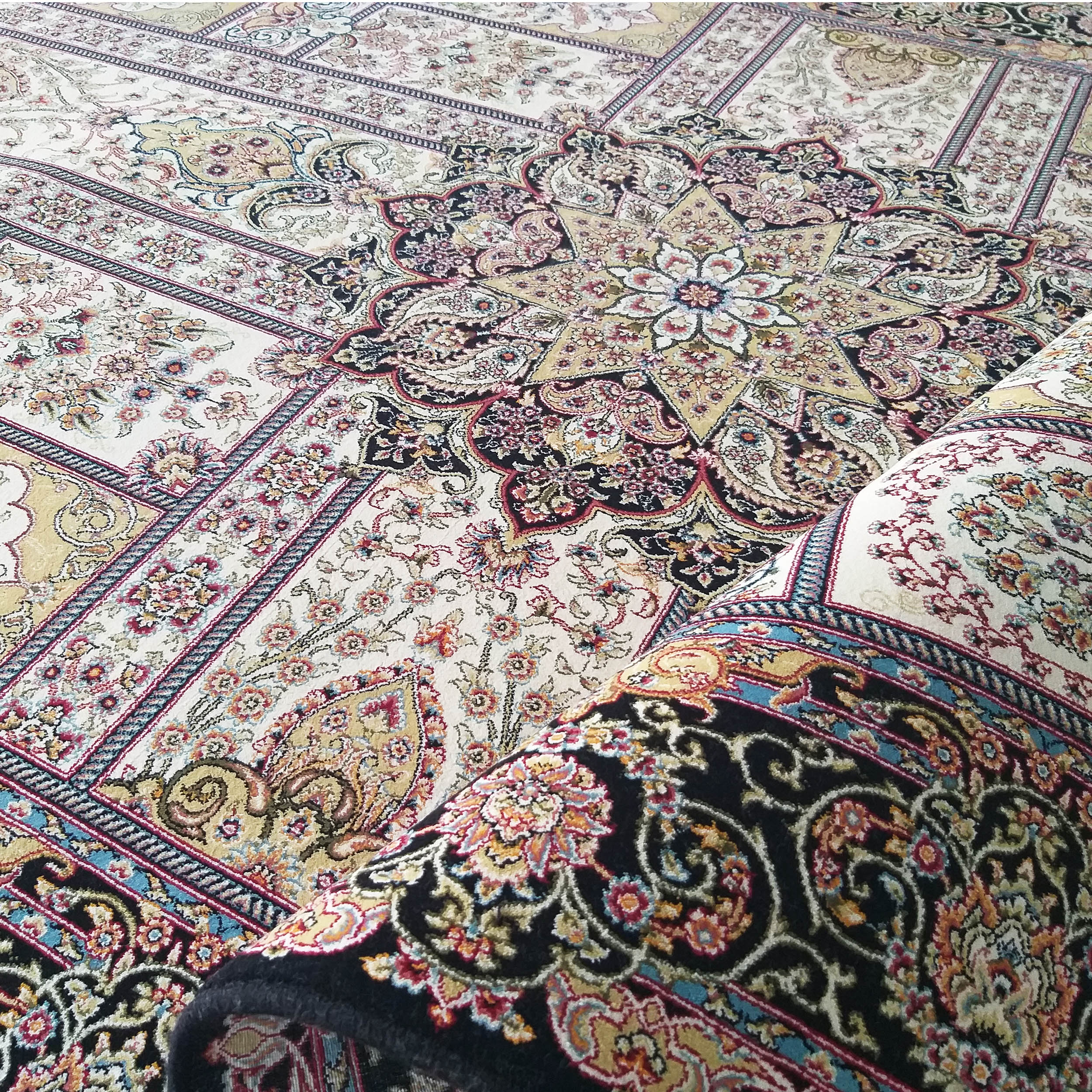 Tapis textile à motifs floraux et médaillons complexes, avec des couleurs crème, noir, or, rouge et bleu, partiellement roulé