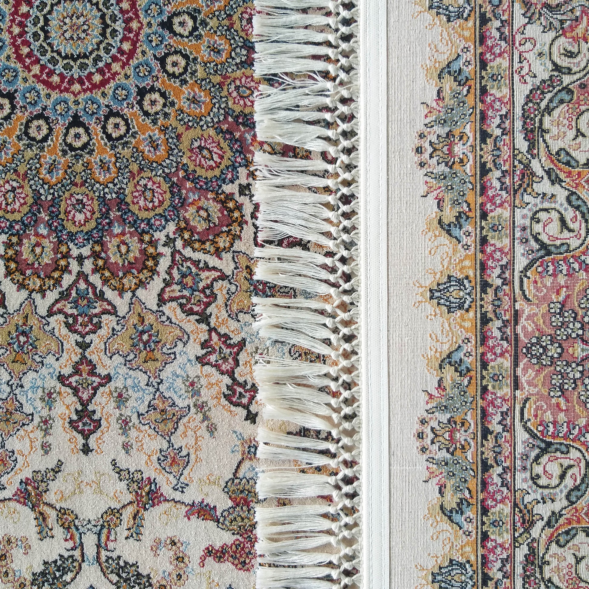 Tapis ornemental aux motifs floraux complexes en rouge, bleu, crème, doré et noir, bordé de franges blanches délicates