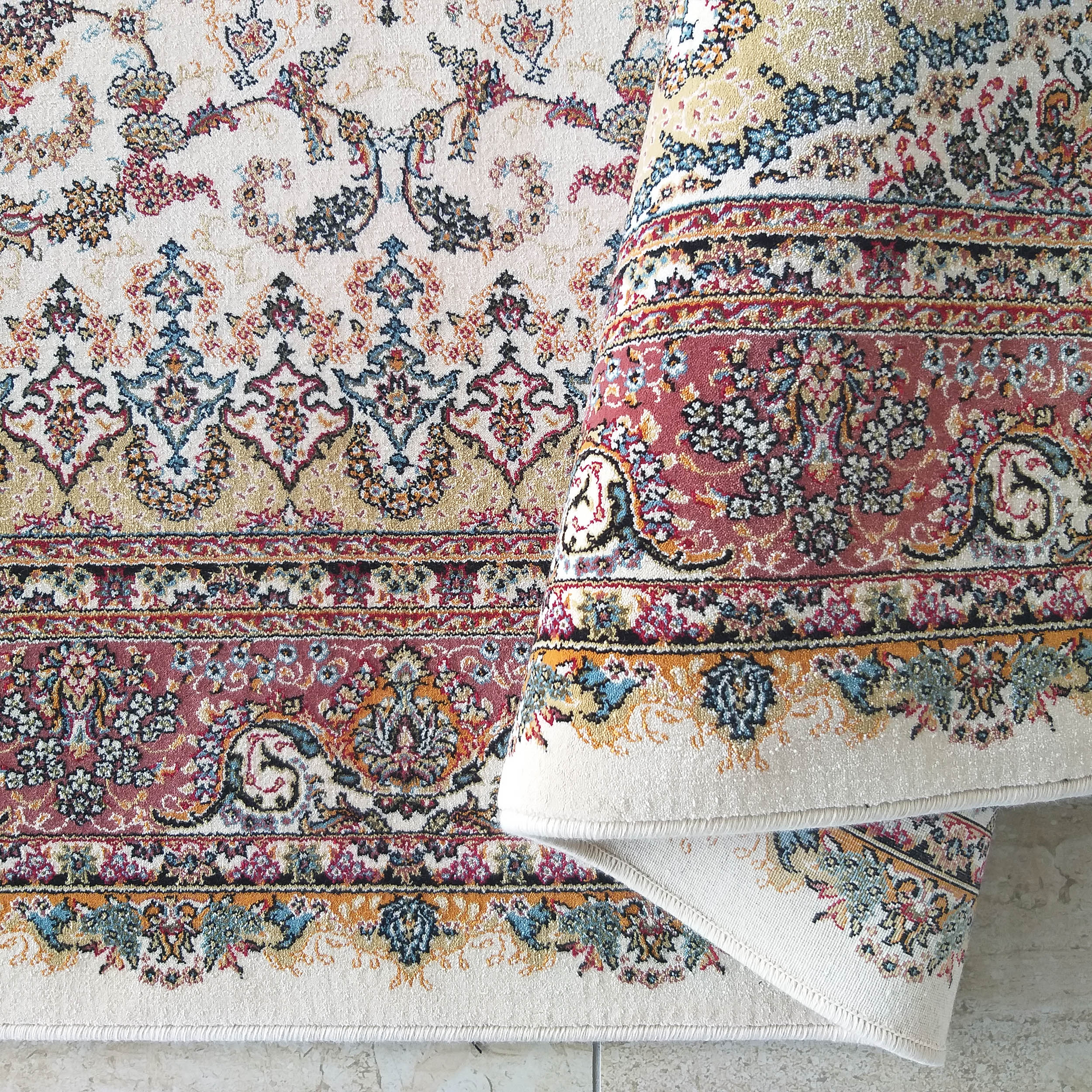 Tapis crème aux motifs traditionnels riches en rouge, bleu, or, noir et vert, avec des bordures détaillées et une partie enroulée