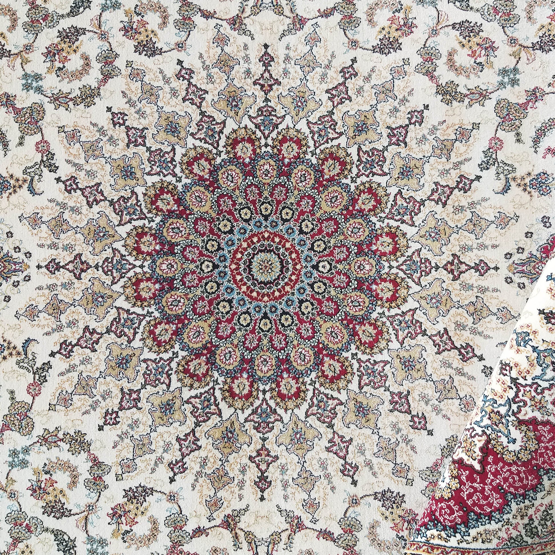 Tapis crème à motifs floraux et arabesques complexes, avec un médaillon central rouge, doré et bleu, et un coin replié