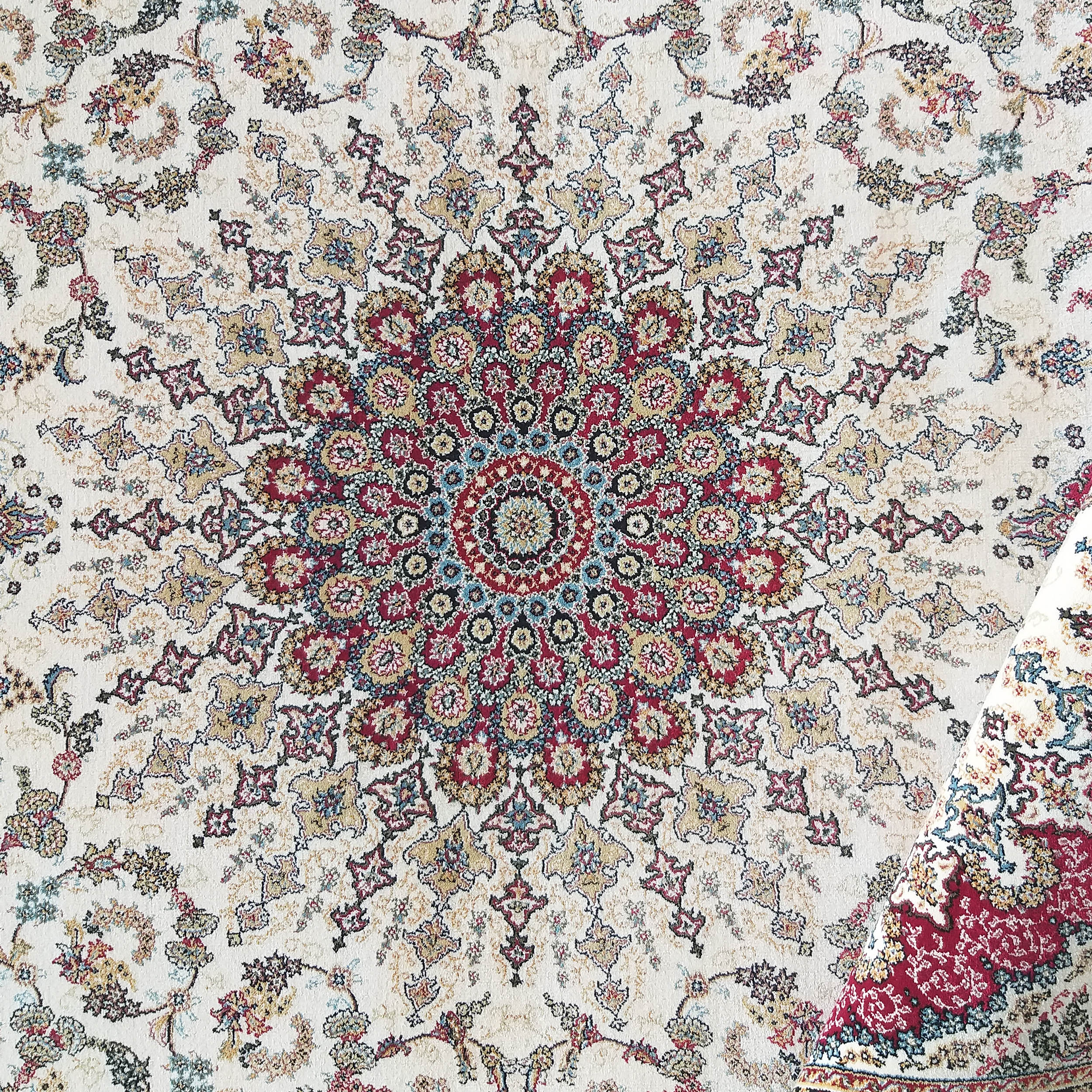 Tapis crème à motifs floraux et arabesques complexes, avec un médaillon central rouge, doré et bleu, et un coin replié