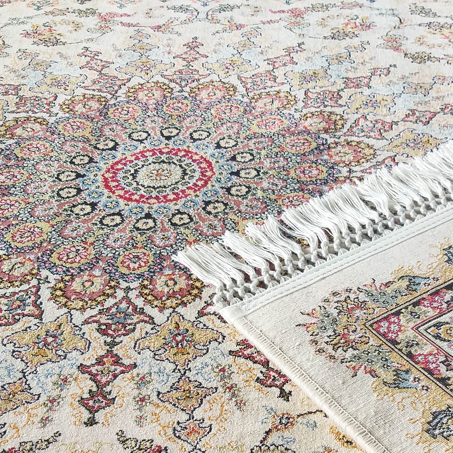 Tapis crème à motifs floraux multicolores rouges, bleus, jaunes et verts. Une section est repliée, montrant ses franges