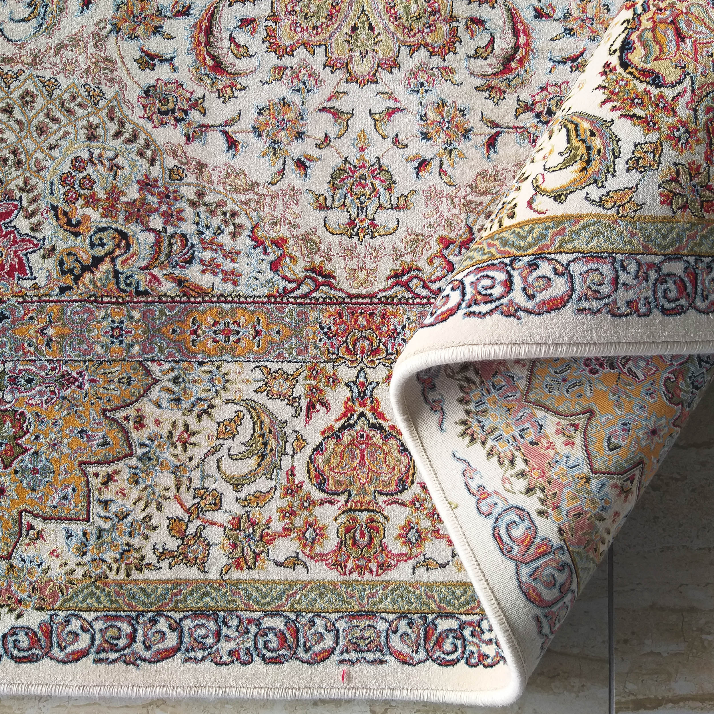 Tapis crème aux riches motifs floraux et arabesques rouges, or et bleus, dont un coin est roulé sur un carrelage clair