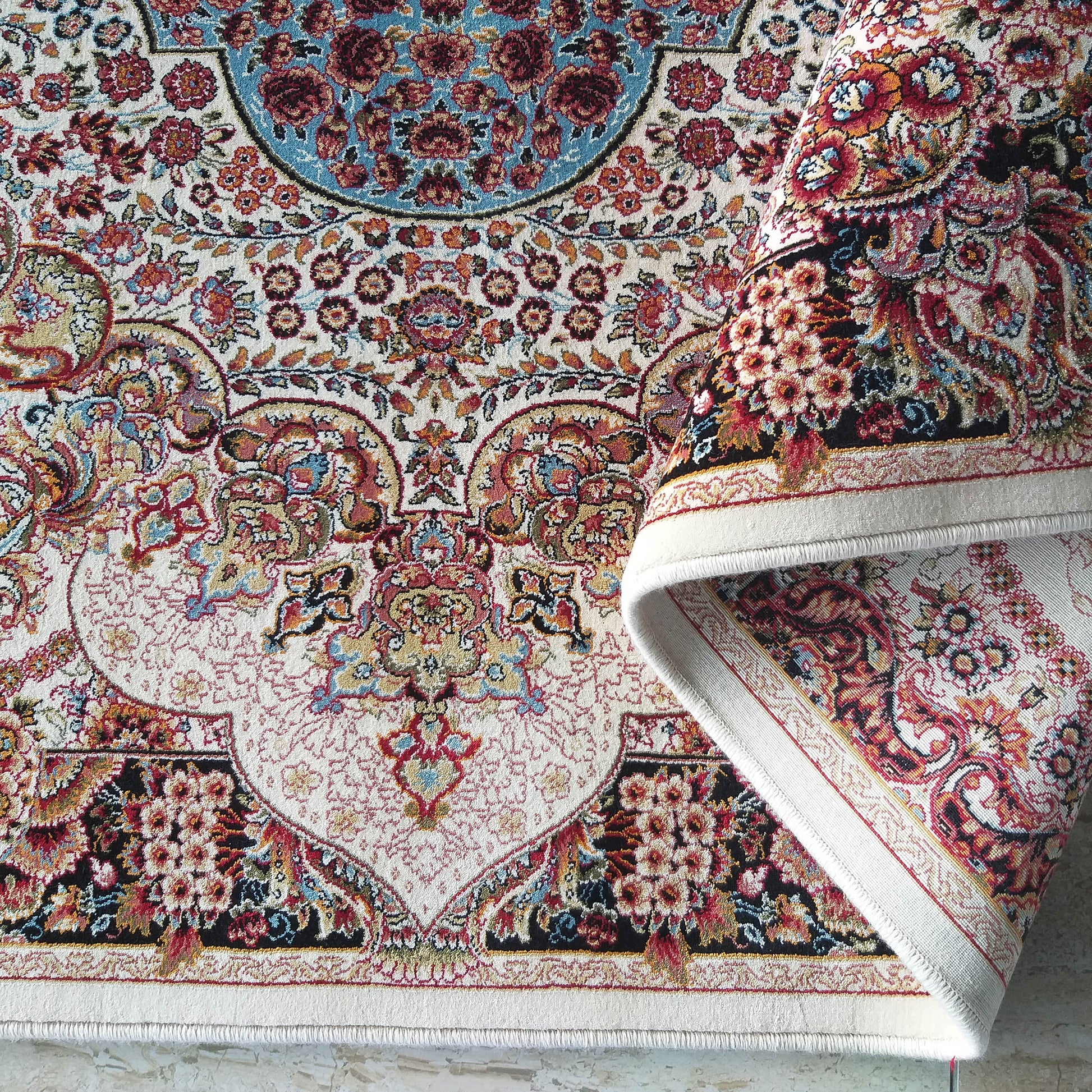 Tapis aux motifs floraux et cachemire complexes, couleurs dominantes rouge, bleu, or et noir sur fond crème clair. Une partie du tapis est roulée