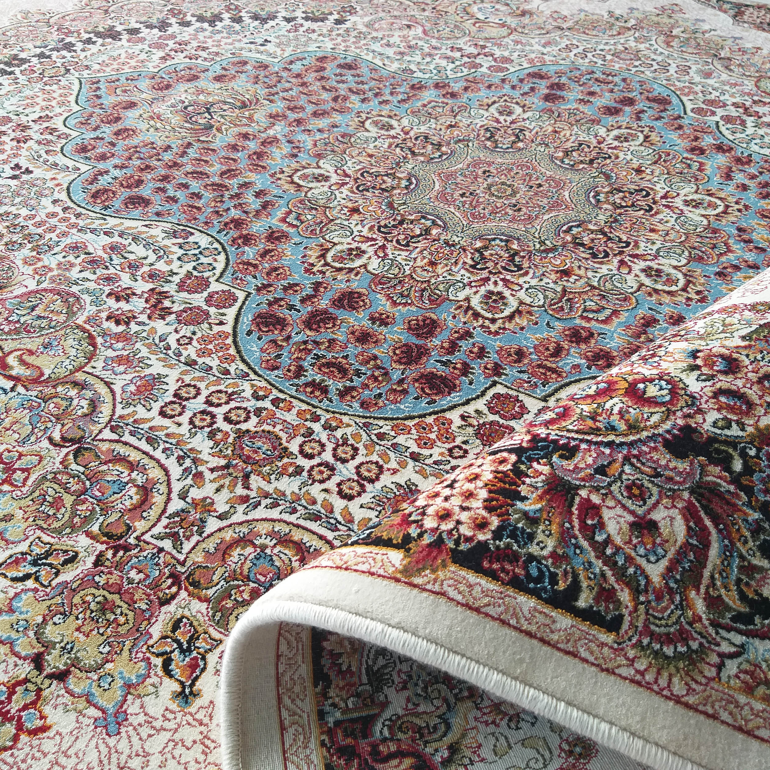 Tapis crème à motifs fleuris et volutes complexes, mêlant le rouge, le bleu, le doré et le noir. Une partie est roulée, révélant les détails du tissage