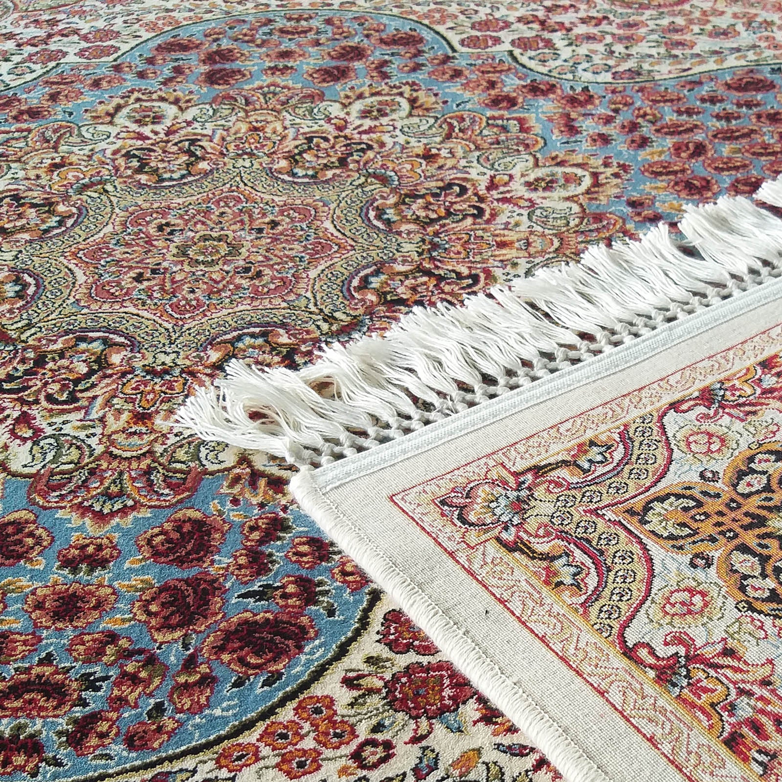 Gros plan sur un tapis aux motifs complexes, floraux et ornementaux, en rouge, crème, bleu et doré. Franges blanches et une étiquette noire