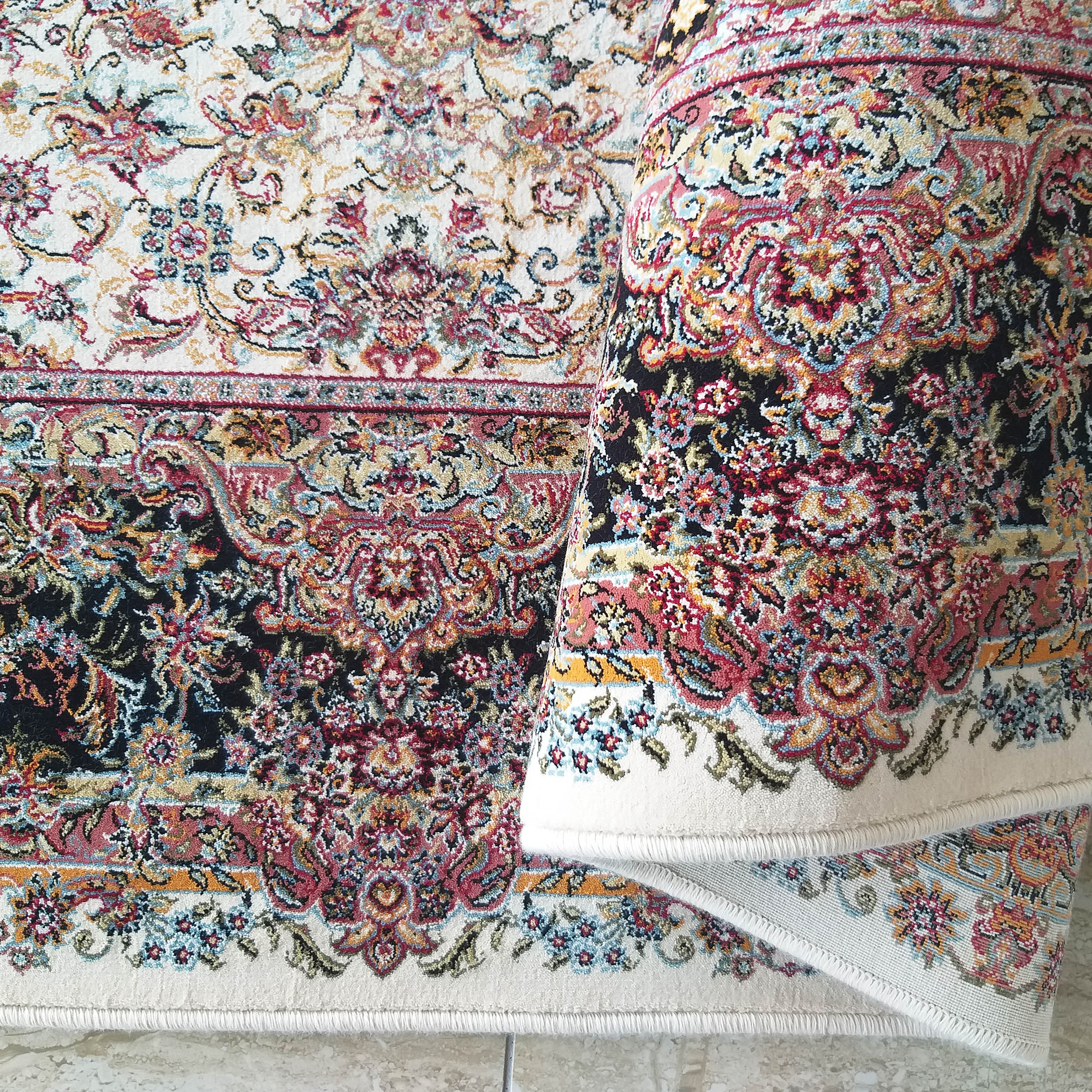 Tapis aux motifs floraux colorés, roses, bleus, jaunes et rouges sur fond crème et noir, une section est enroulée
