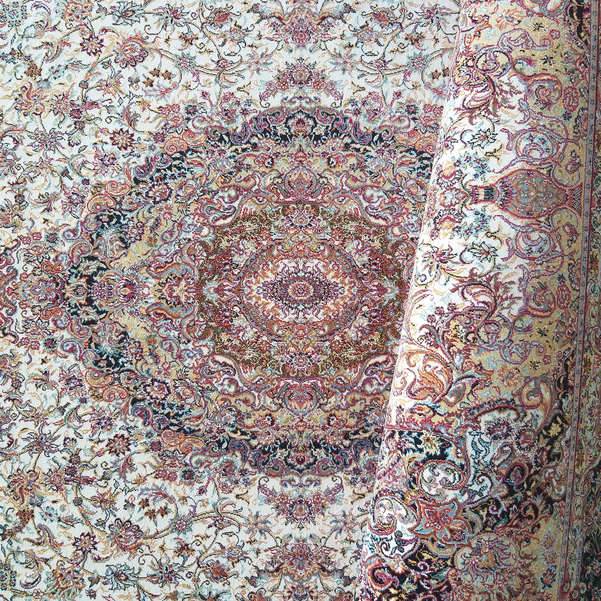 Tapis à fond écru avec motifs floraux et un grand médaillon central aux couleurs rouge, or, bleu et noir, en partie roulé