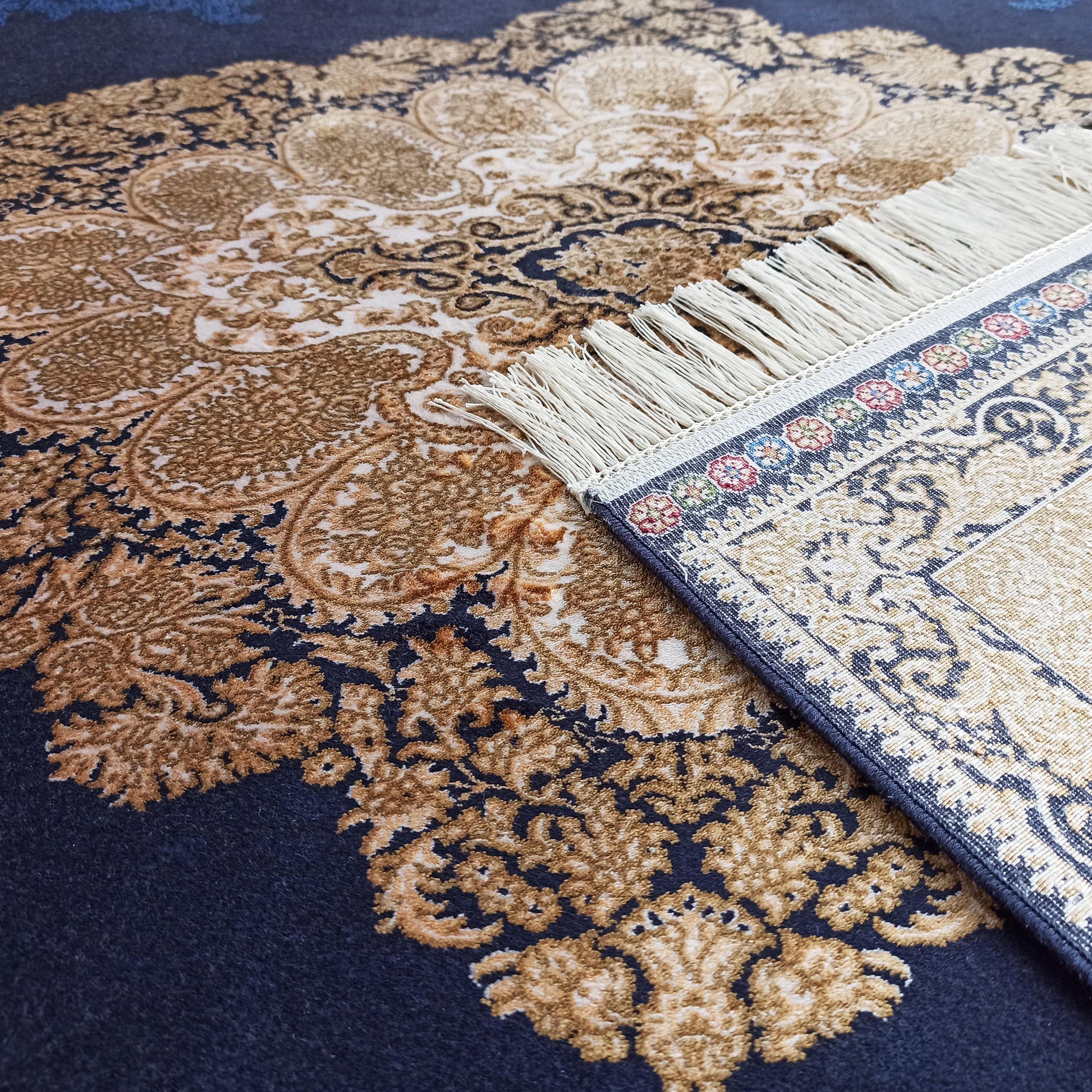 Tapis aux motifs ornés bleu marine et or, avec une bordure à franges claires et des détails floraux colorés