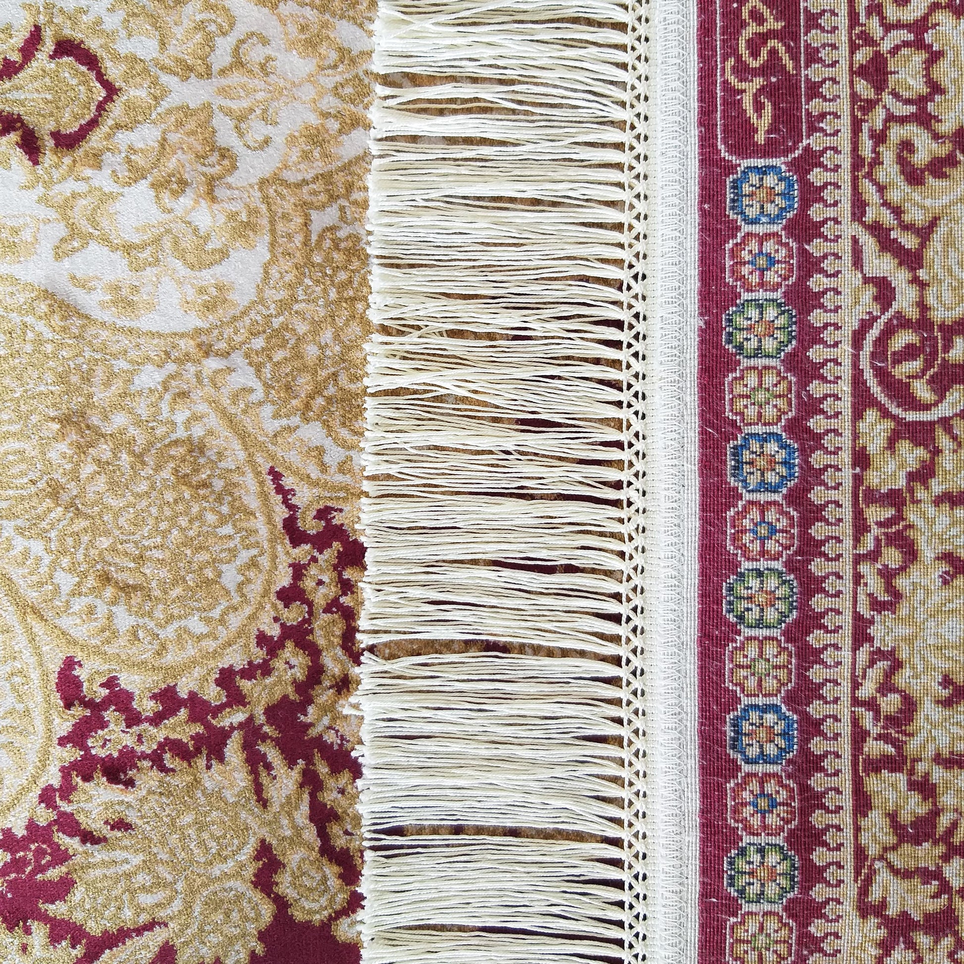 Tapis décoré de motifs floraux et géométriques en bordeaux, doré et crème, avec une bande de franges blanches