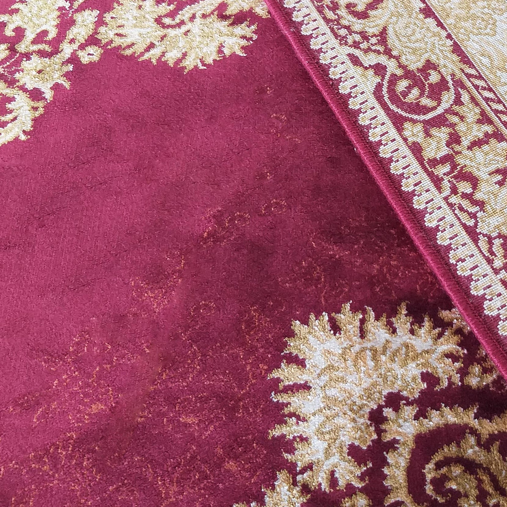 Gros plan sur un tapis ornemental aux motifs floraux dorés sur fond rouge bordeaux, avec une bordure crème et des franges
