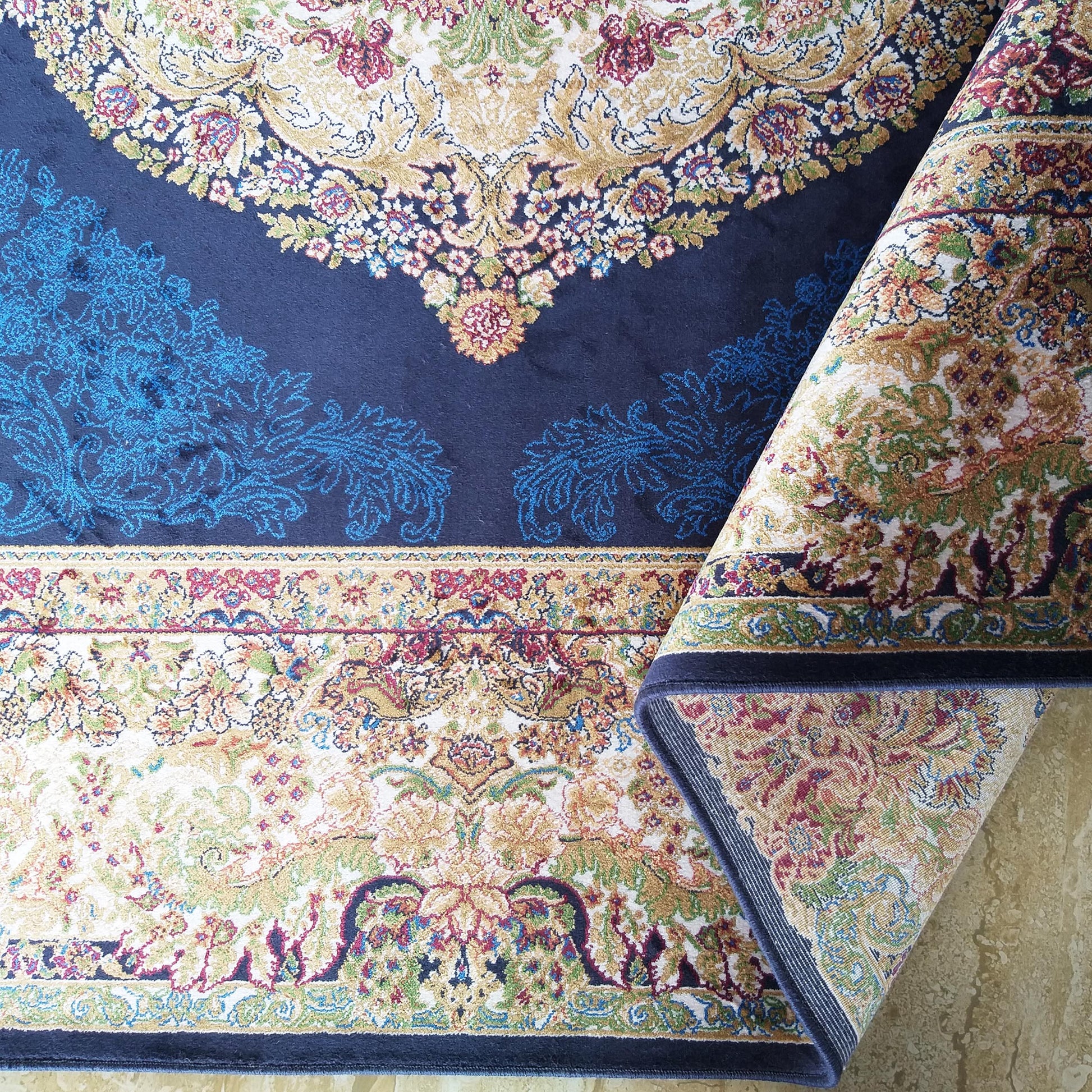 Tapis aux motifs floraux et ornés dorés, rouges, bleus et verts sur fond bleu foncé, partiellement roulé, posé sur un carrelage clair