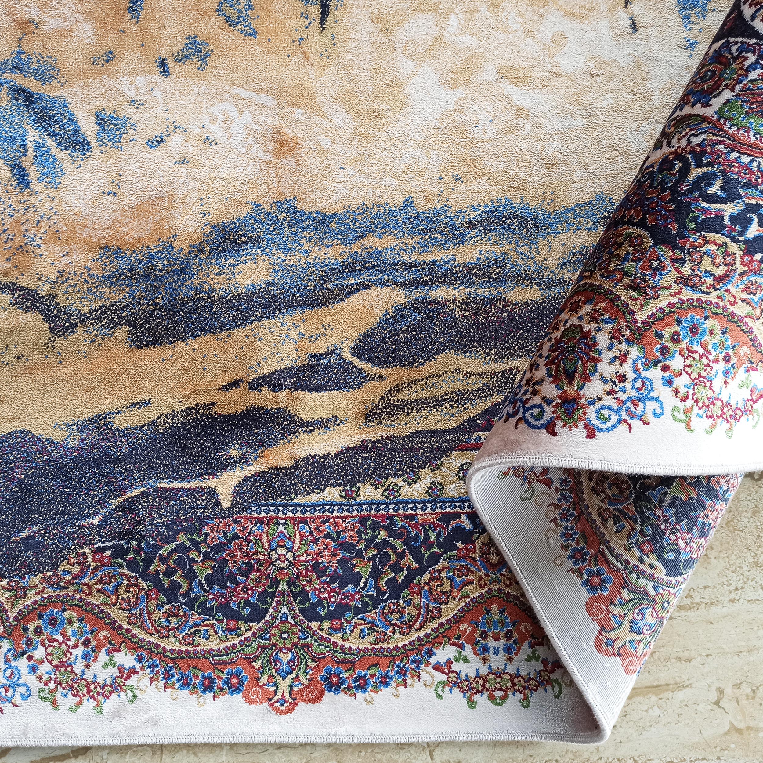 Tapis aux motifs abstraits beige, bleu foncé et clair. Une partie roulée révèle une bordure florale multicolore