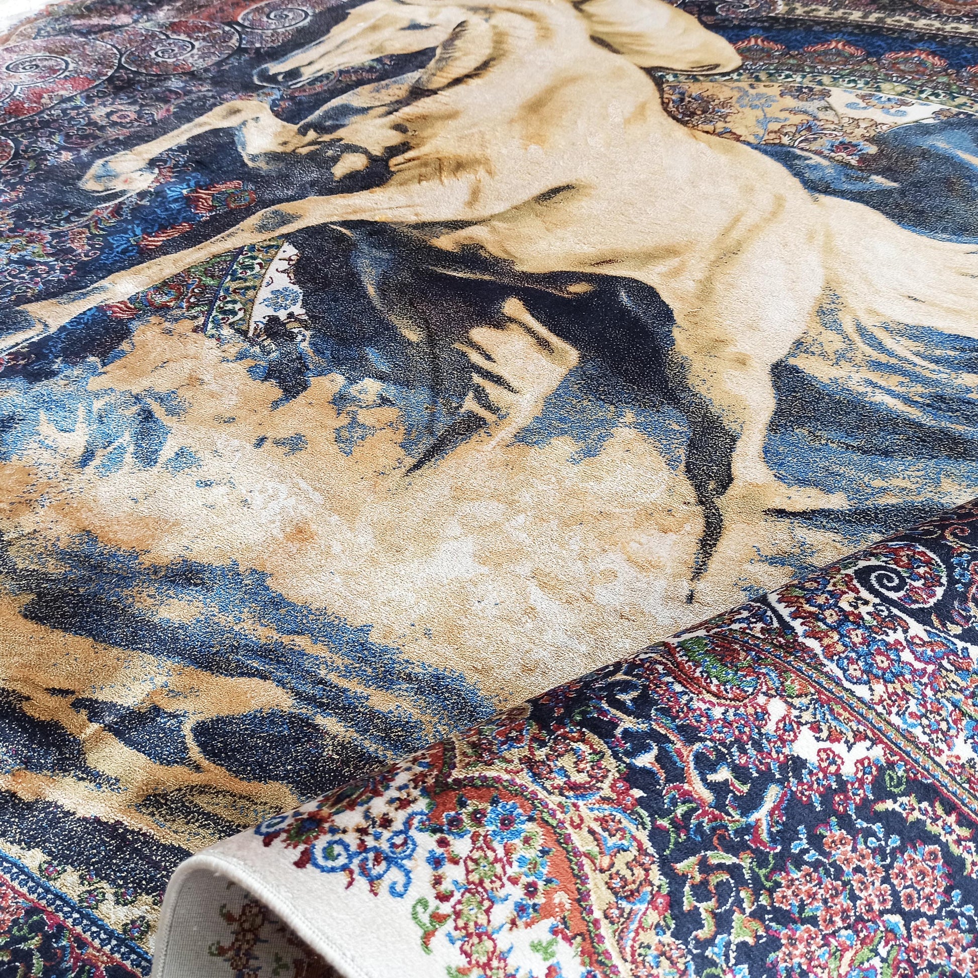 Tapis montrant un cheval beige cabré sur fond bleu et ocre, avec une partie enroulée révélant un motif floral coloré