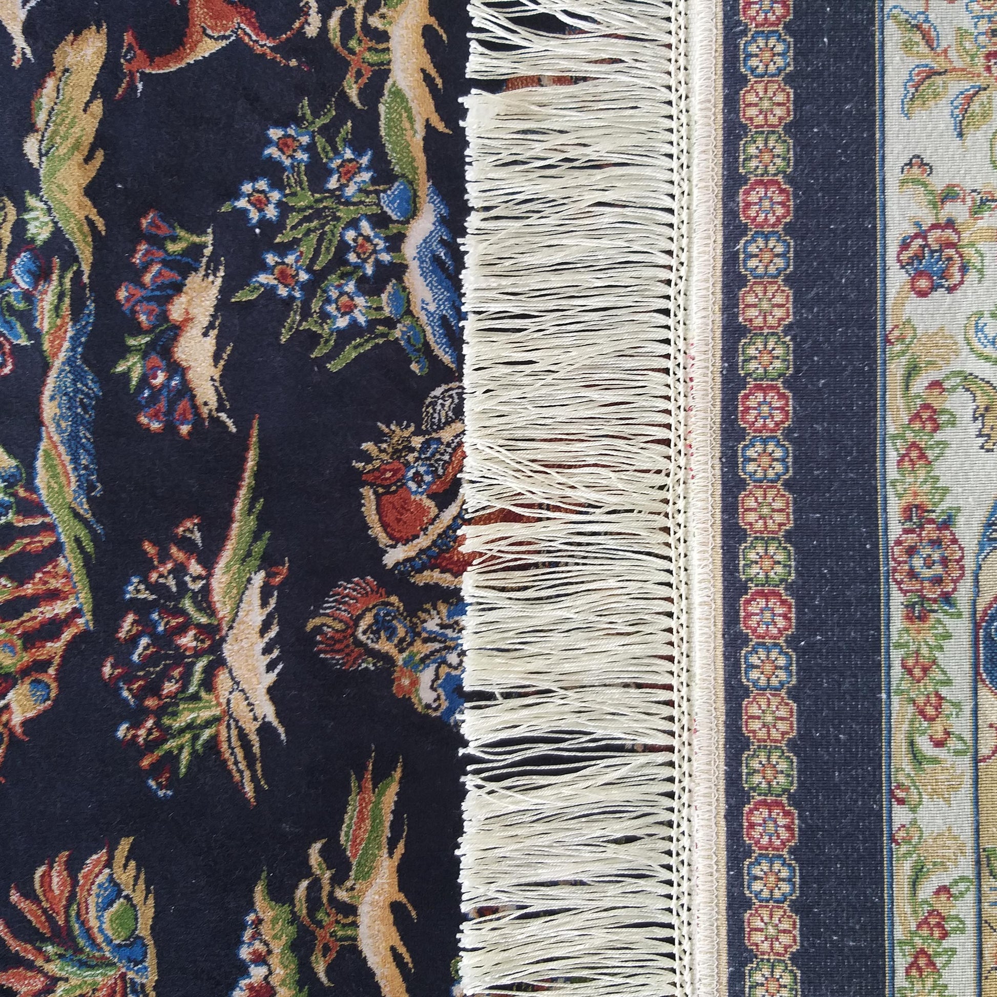 Tapis coloré à motifs floraux et animaux fond bleu foncé à gauche bord à franges et bande à motifs circulaires à droite sur fond clair