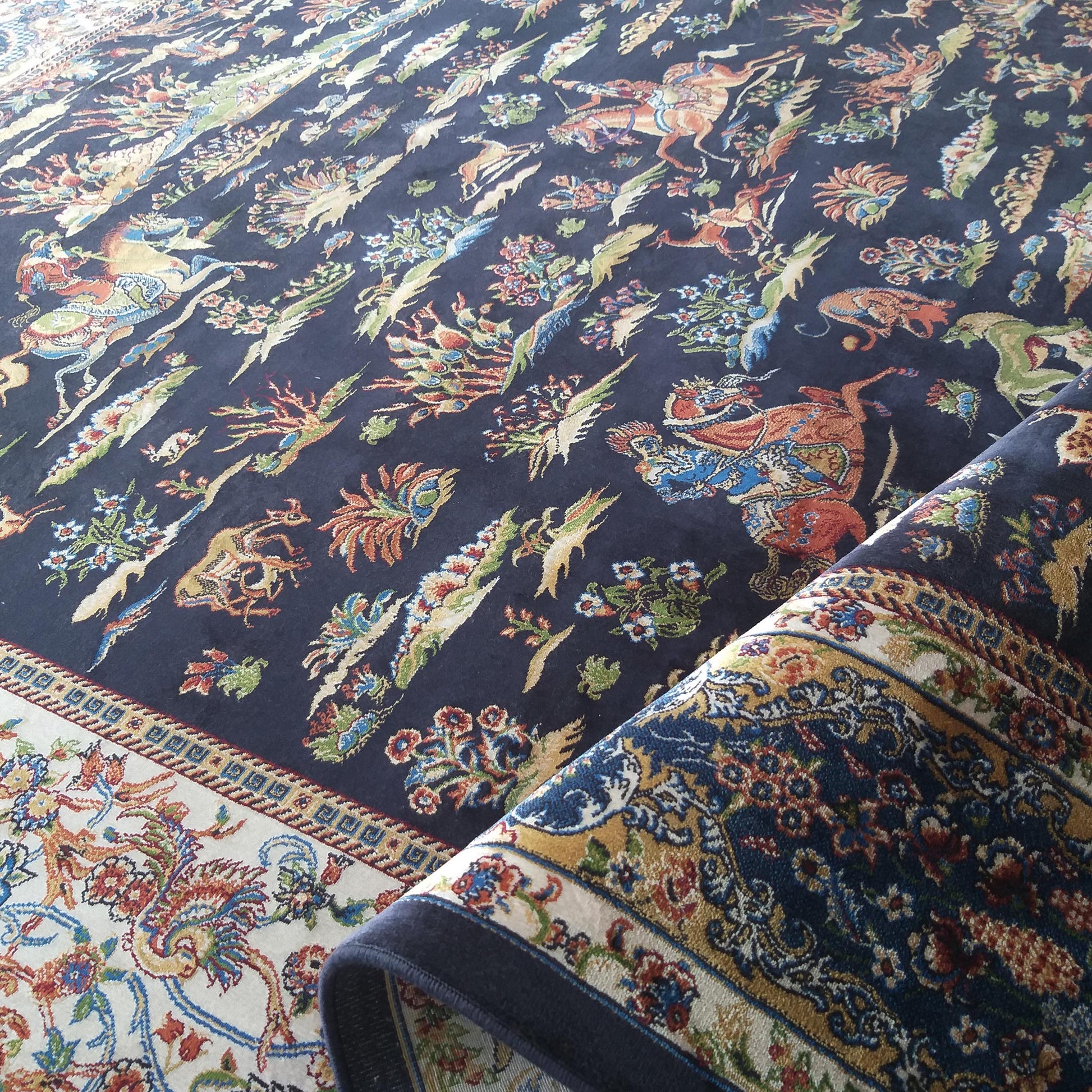 Tapis tissé bleu nuit à motifs floraux et animaliers colorés, avec une bordure beige ornée, partiellement roulé