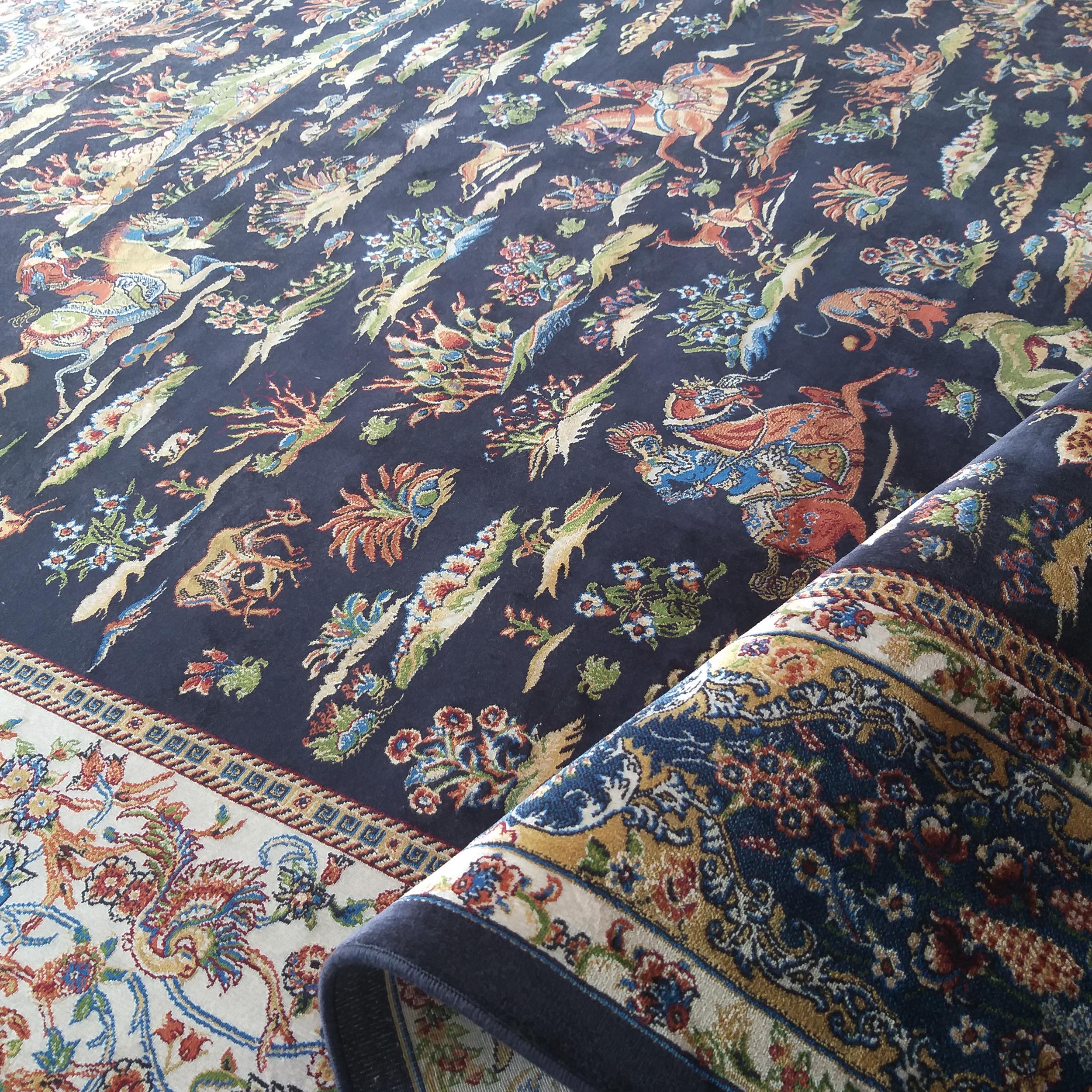 Tapis tissé bleu nuit à motifs floraux et animaliers colorés, avec une bordure beige ornée, partiellement roulé
