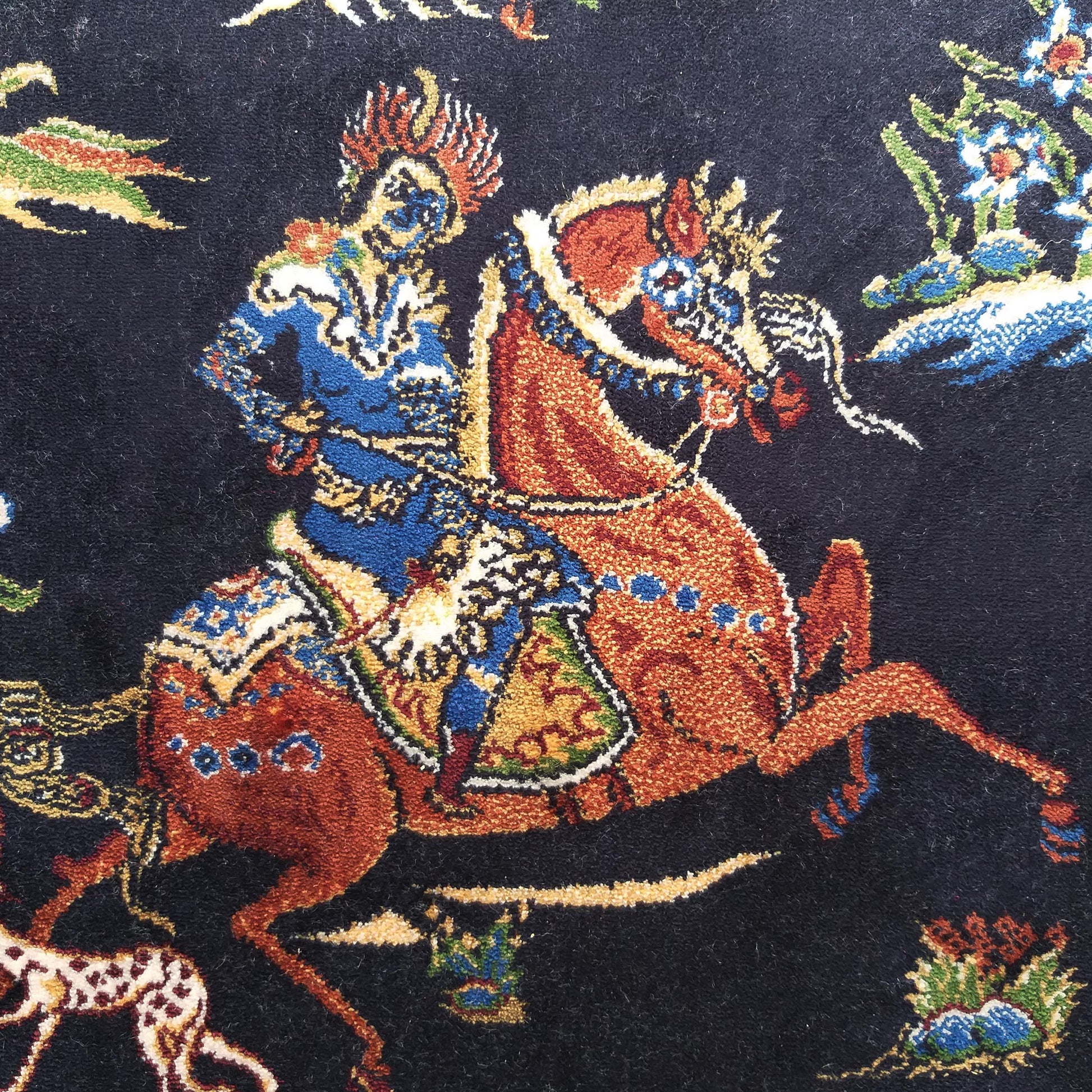 Tapis foncé avec un cavalier en habit bleu et or sur un cheval roux décoré, entouré de motifs floraux colorés