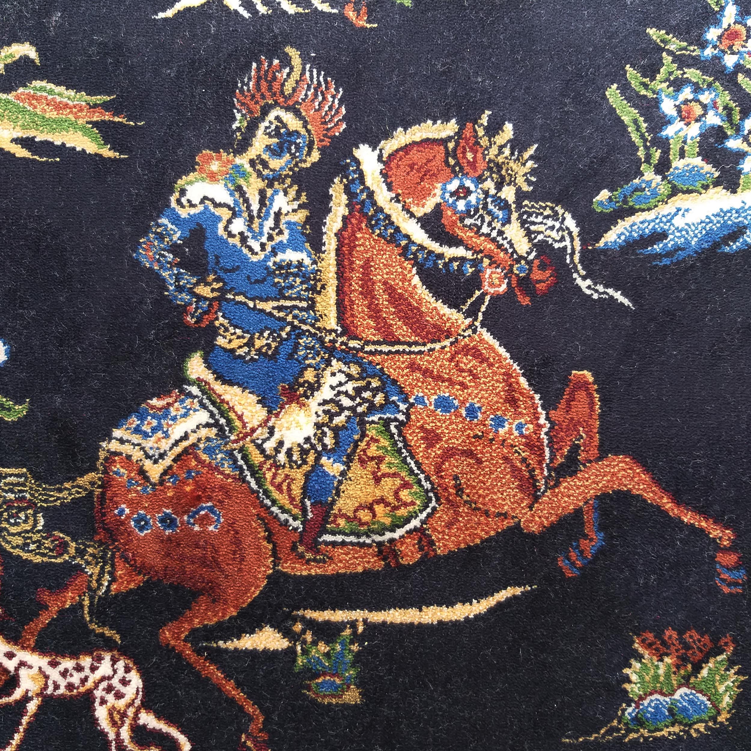 Tapis foncé avec un cavalier en habit bleu et or sur un cheval roux décoré, entouré de motifs floraux colorés
