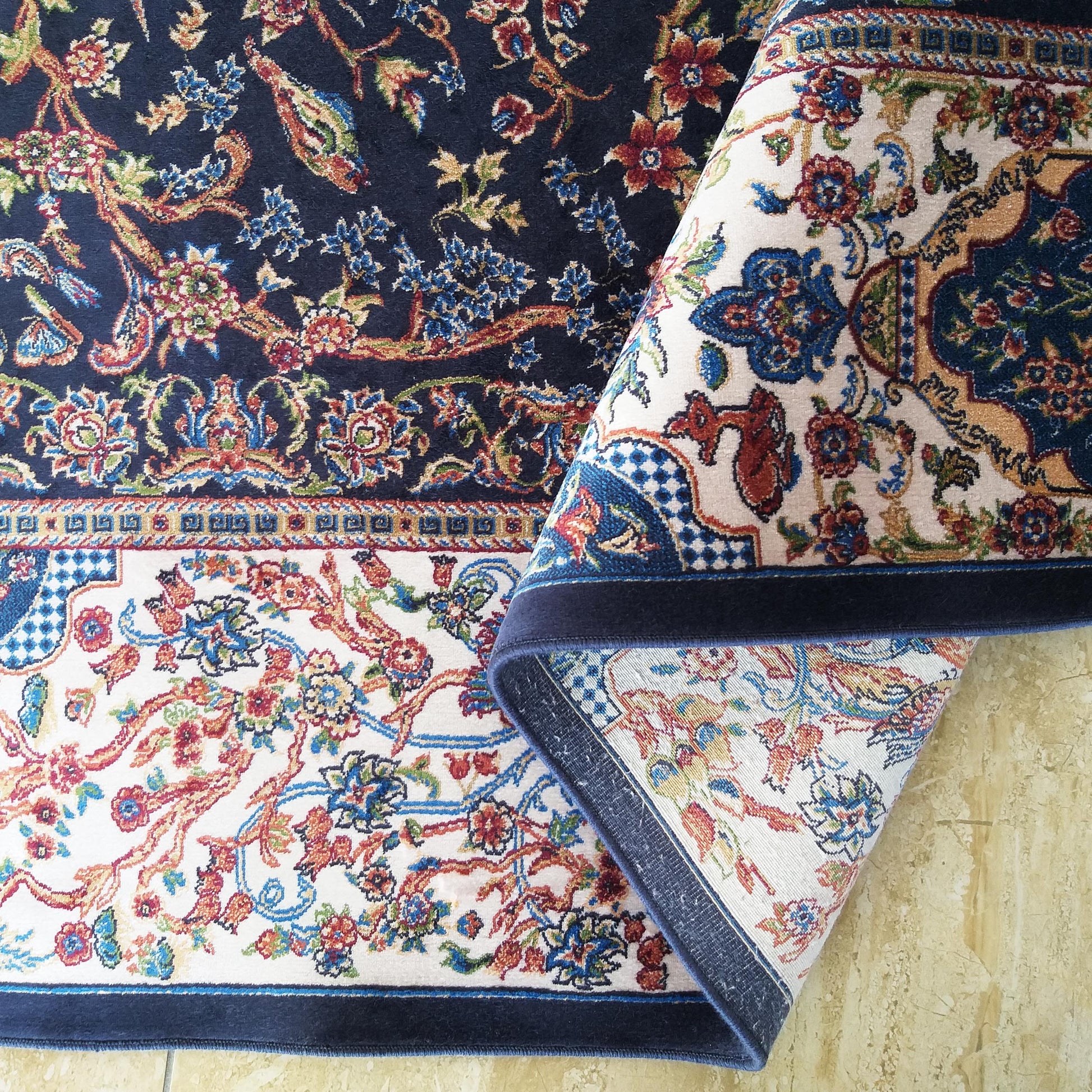 Tapis oriental bleu marine à motifs floraux complexes et arabesques multicolores, partiellement roulé révélant un fond crème