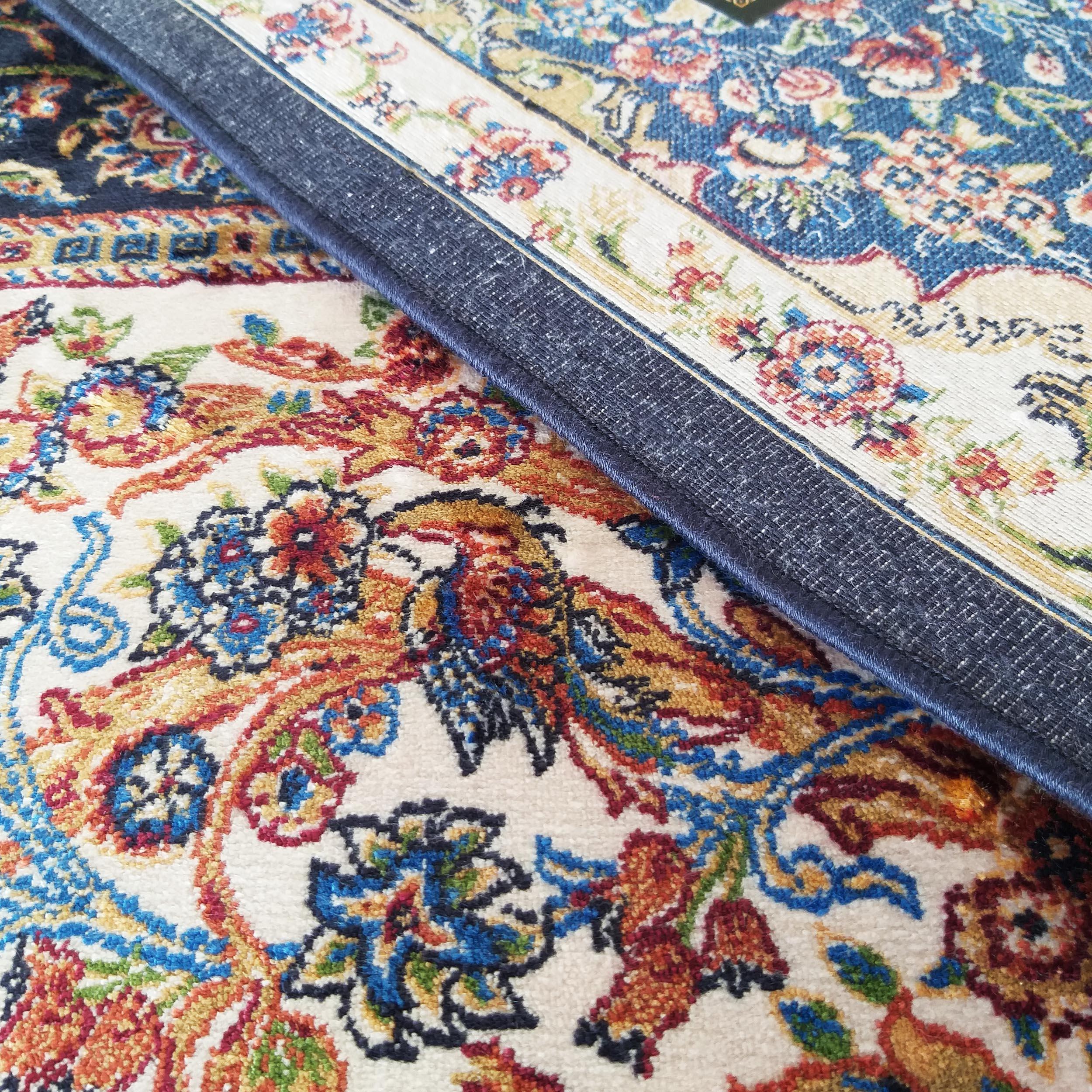 Un tapis blanc cassé orné de motifs floraux et d'oiseaux polychromes, partiellement recouvert par un tapis bleu à motifs