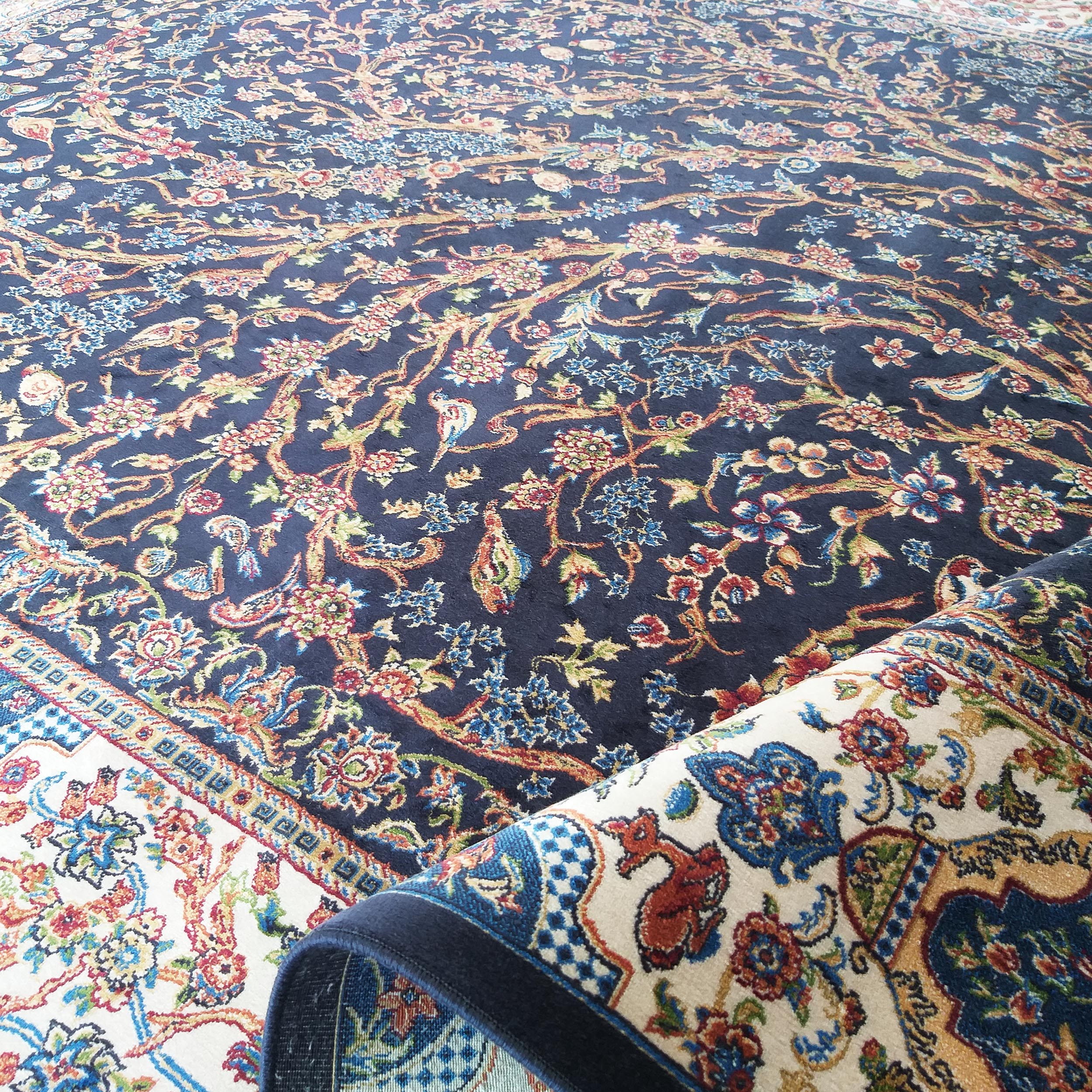 Grand tapis bleu foncé richement orné de motifs floraux et d'oiseaux, avec une section roulée montrant une bordure crème