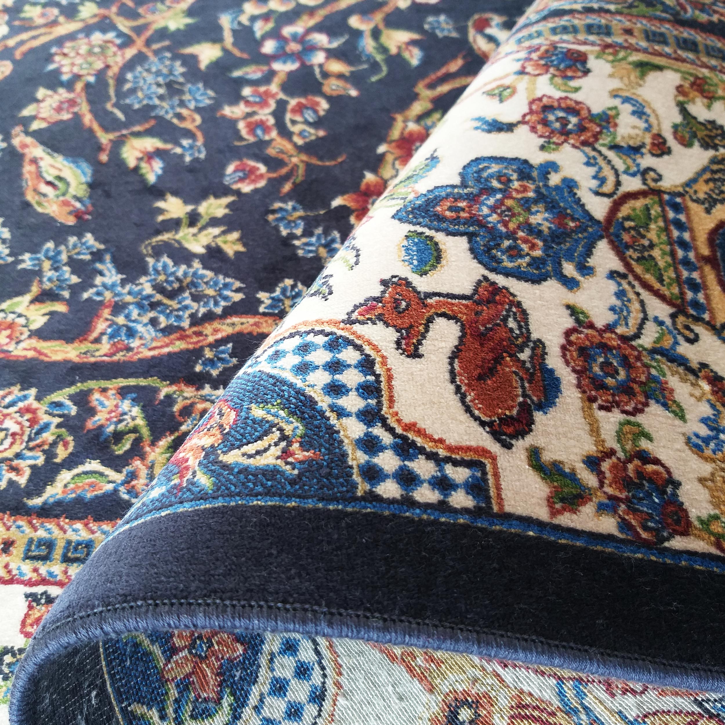Tapis aux motifs floraux et arabesques multicolores, un coin est replié, révélant une bordure crème et le dos bleu foncé