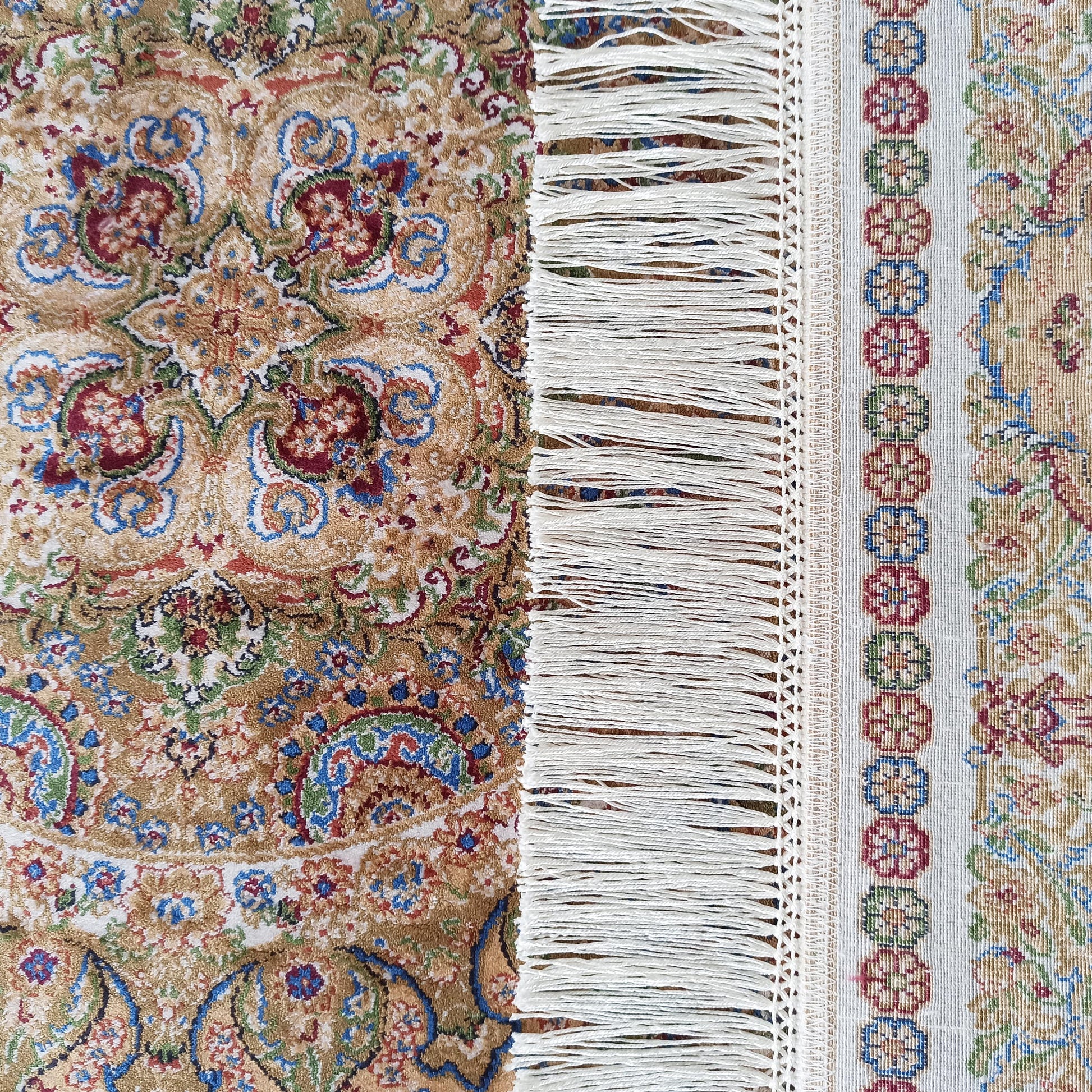 Gros plan sur un tapis beige orné de motifs floraux et arabesques rouges, bleues et vertes, avec une bordure à franges blanches