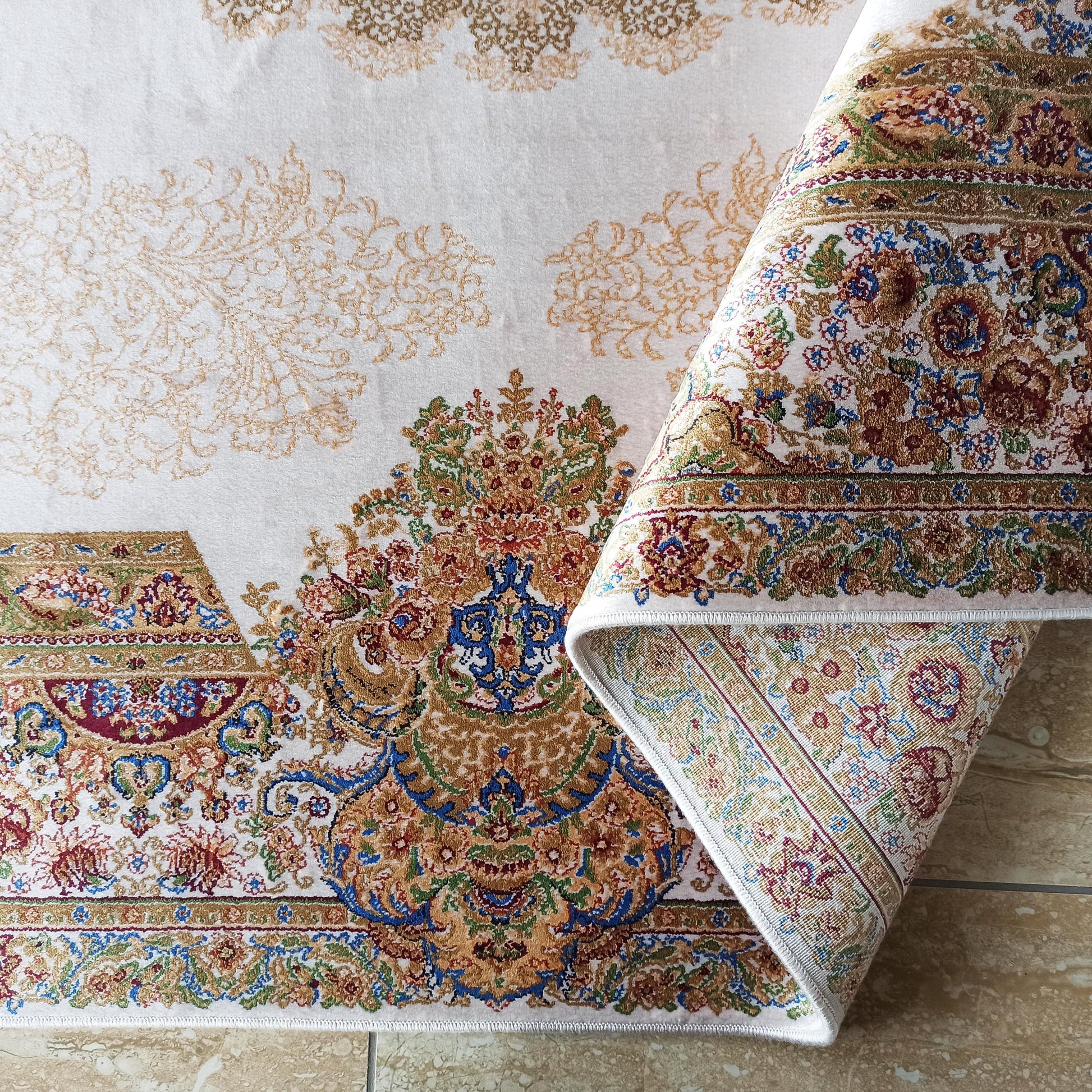 Tapis crème à motifs floraux dorés, dont une section enroulée révèle un design multicolore riche en bordeaux, bleu et vert