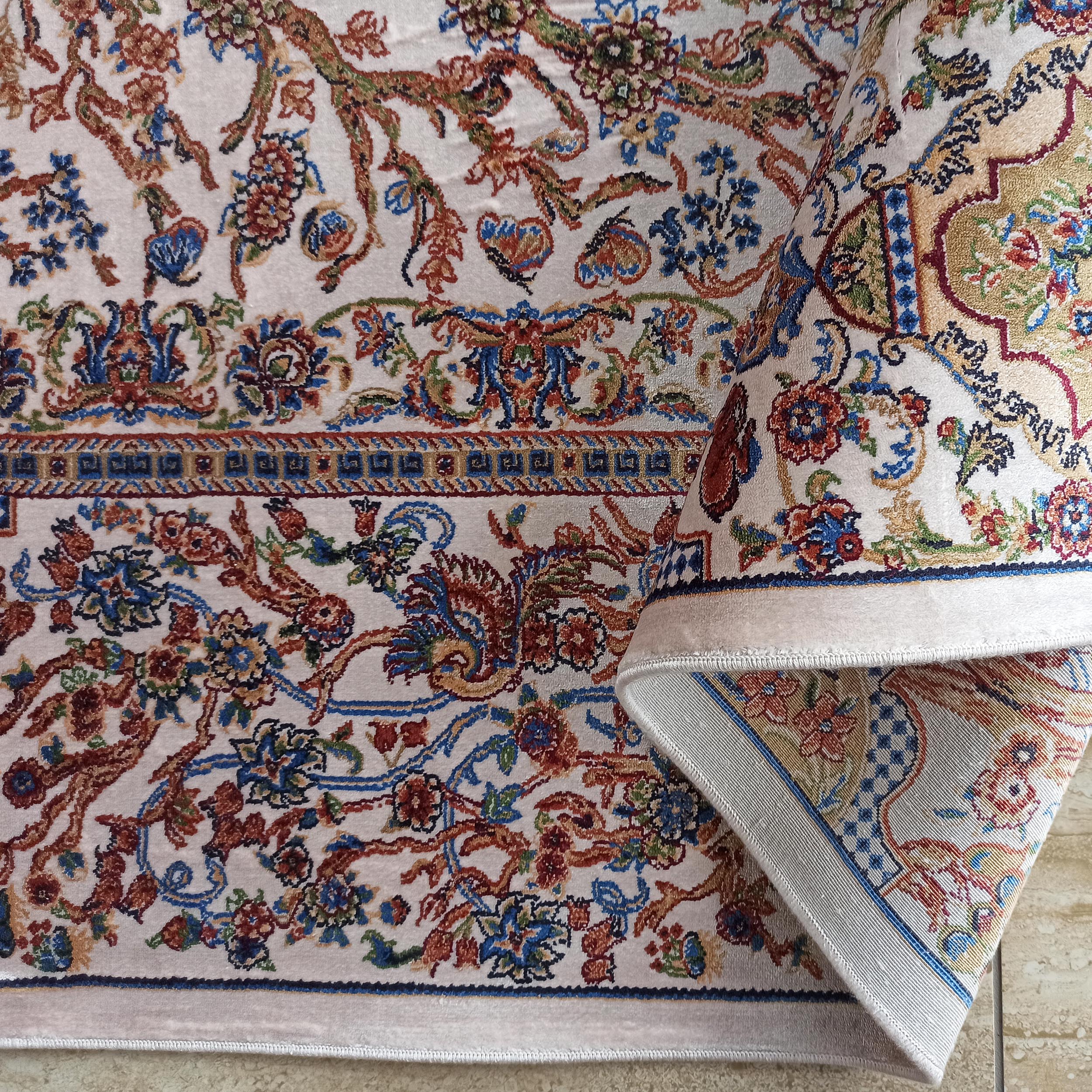 Tapis crème à motifs floraux complexes et entrelacés de bleu, rouge, vert et or. Le tapis est partiellement enroulé
