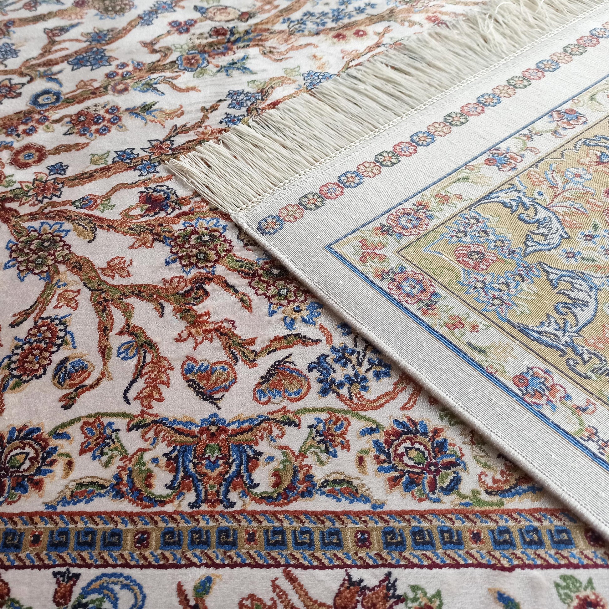 Tapis aux motifs floraux et végétaux complexes en bleu, rouge, vert et ocre sur fond crème, avec une bordure ornée et une frange