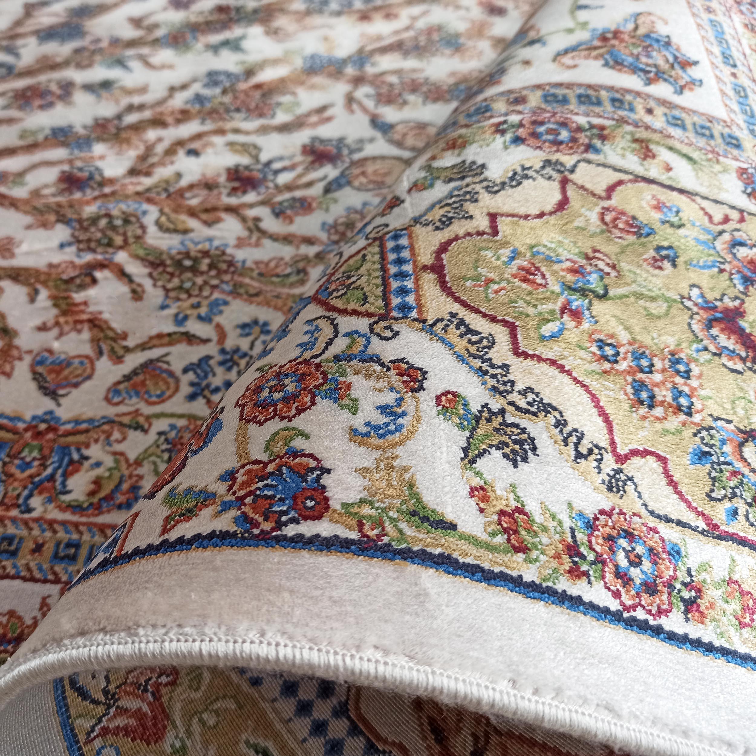 Tapis crème partiellement enroulé, orné de motifs floraux et arabesques multicolores en bleu, rouge, vert et doré
