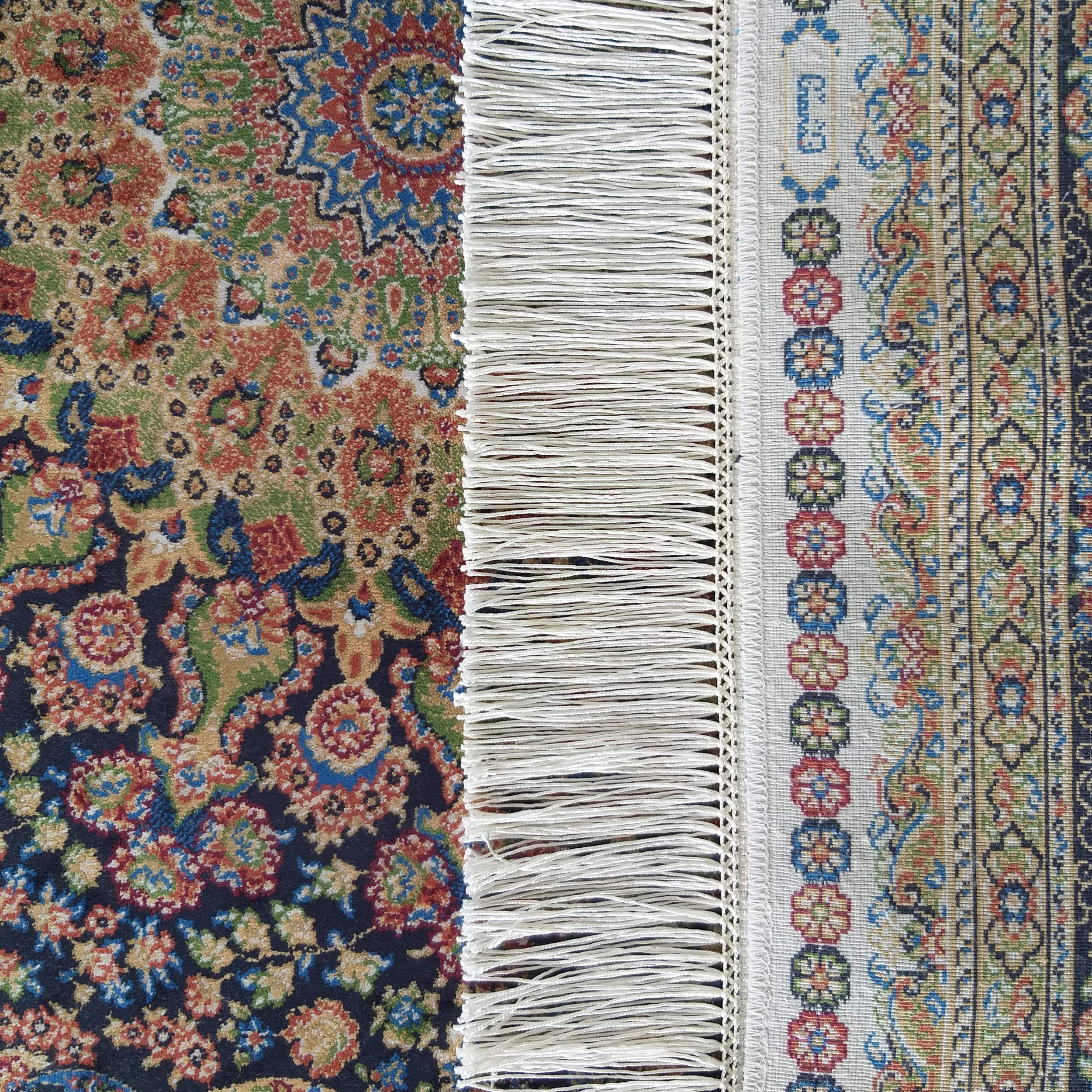 Tapis coloré avec motifs floraux complexes. À gauche, un fond bleu foncé et à droite, une bordure claire avec des franges blanches au centre