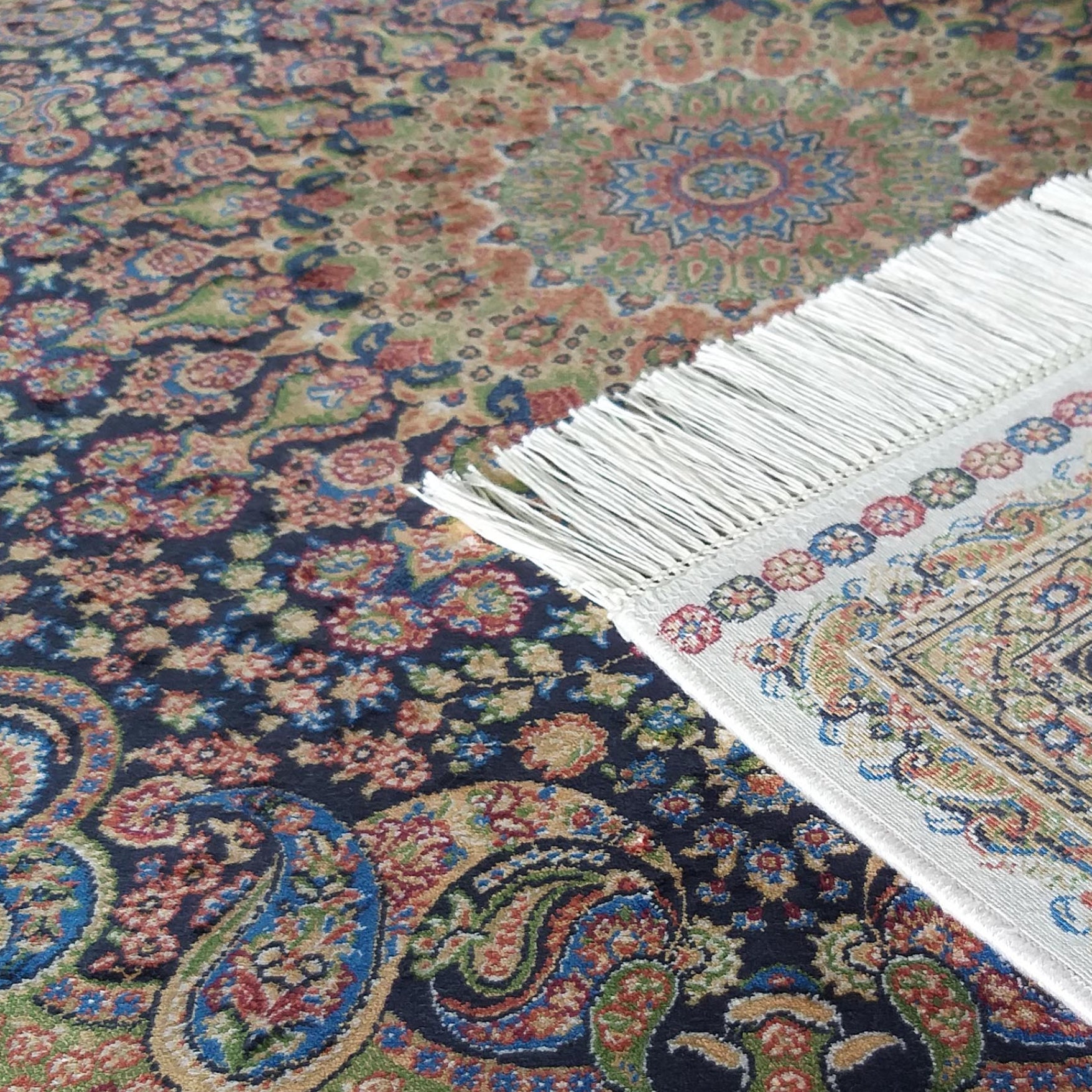 Gros plan sur un tapis sombre à motifs floraux et cachemires multicolores, avec le coin d'un tapis clair à franges blanches soulevé