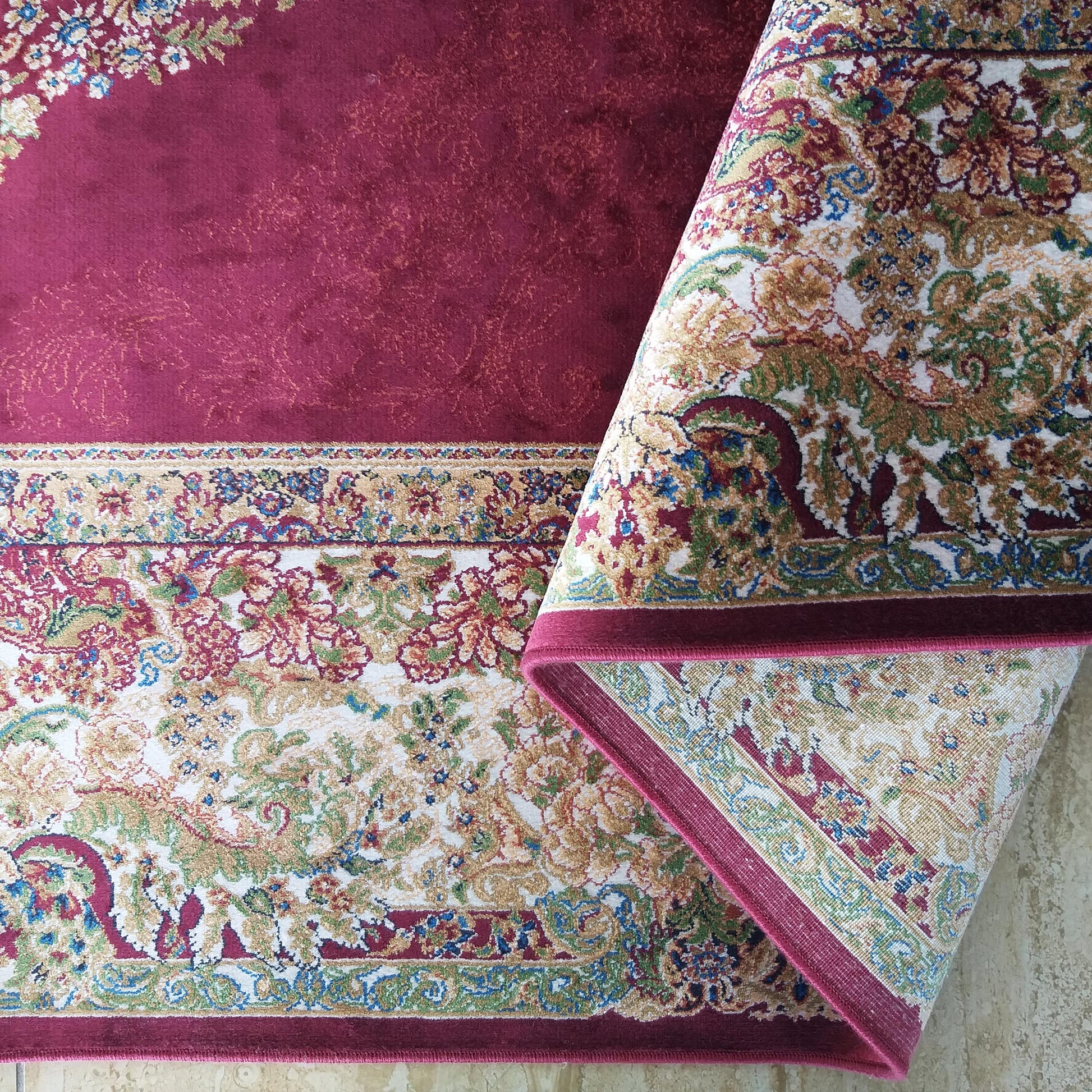 Tapis bordeaux avec des motifs fleuris or, verts et bleus sur fond crème, partiellement enroulé sur la droite