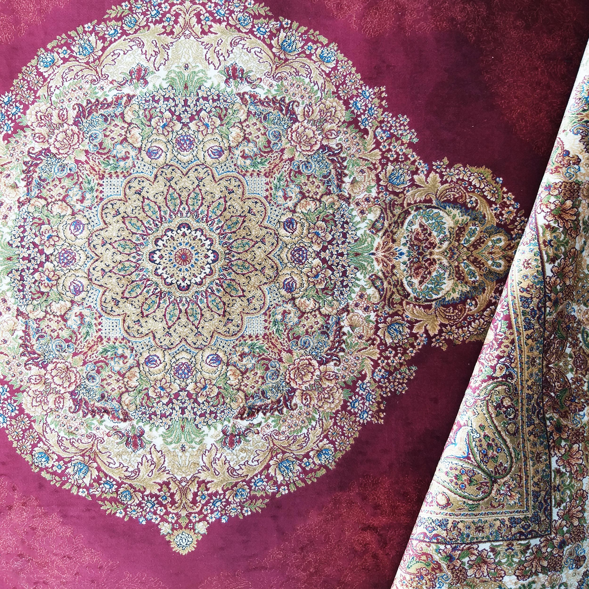 Tapis bordeaux avec un grand médaillon central et de riches motifs floraux dorés, bleus et verts. Une partie du tapis est roulée