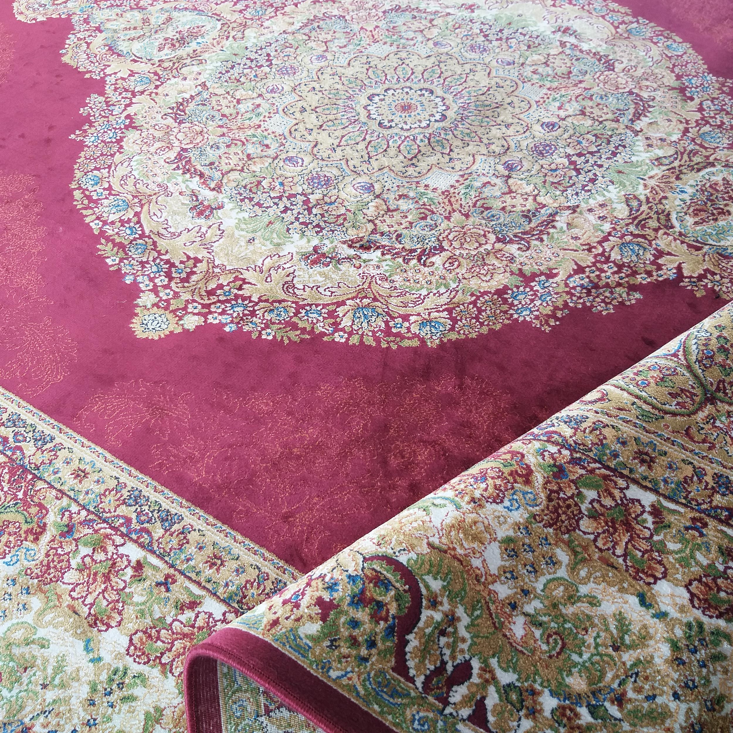 Tapis rouge bordeaux avec un grand médaillon central et des motifs floraux complexes beige, or, vert et bleu, partiellement roulé