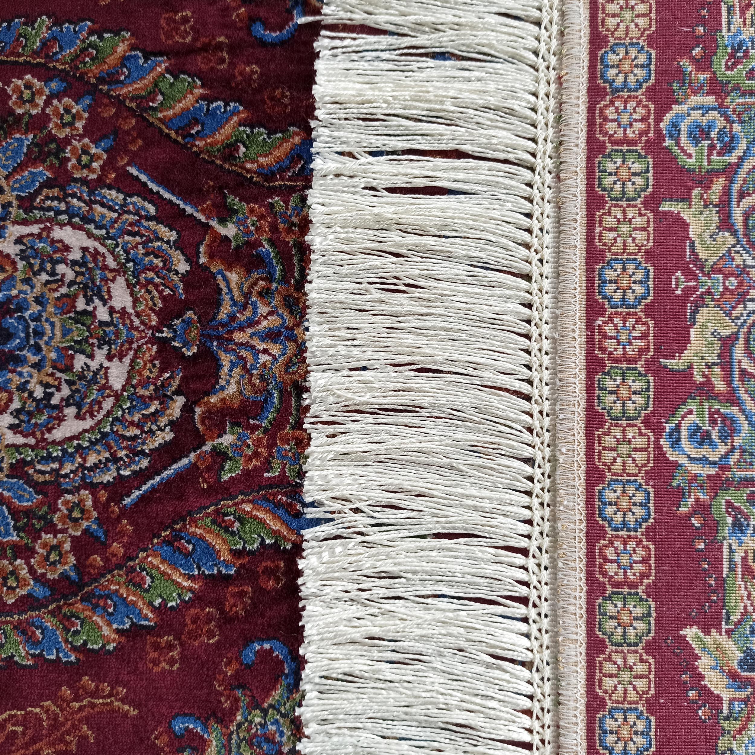 Un tapis bordeaux richement orné de motifs floraux bleus, verts et crème. Il présente des franges blanches et une bordure à motifs géométriques multicolores