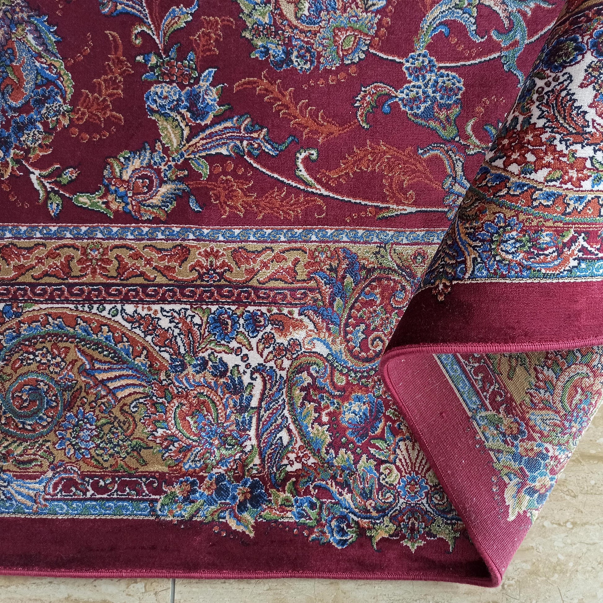 Tapis bordeaux à motifs floraux et cachemire multicolores, partiellement roulé, montrant des bordures détaillées et une texture dense