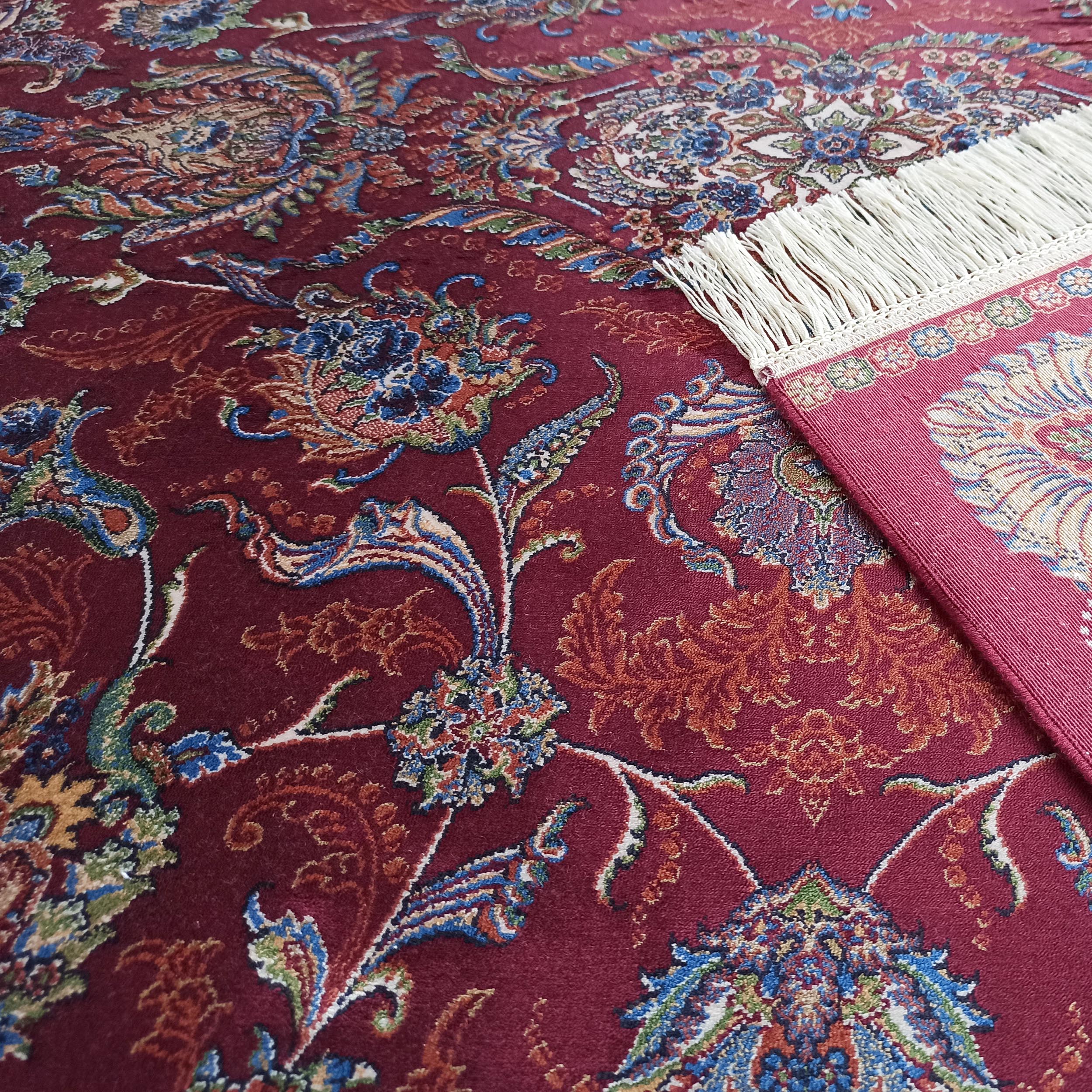 Tapis bordeaux orné de motifs floraux et cachemire bleus, verts et orangés. Une bordure à franges blanches est visible