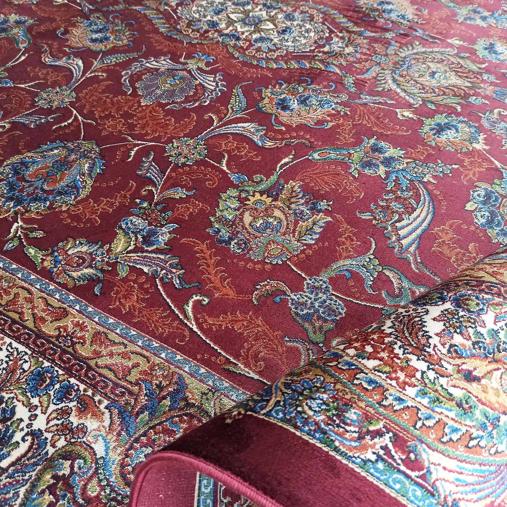 Tapis rouge bordeaux aux motifs floraux et arabesques multicolores bleu, vert, ocre et blanc, partiellement roulé
