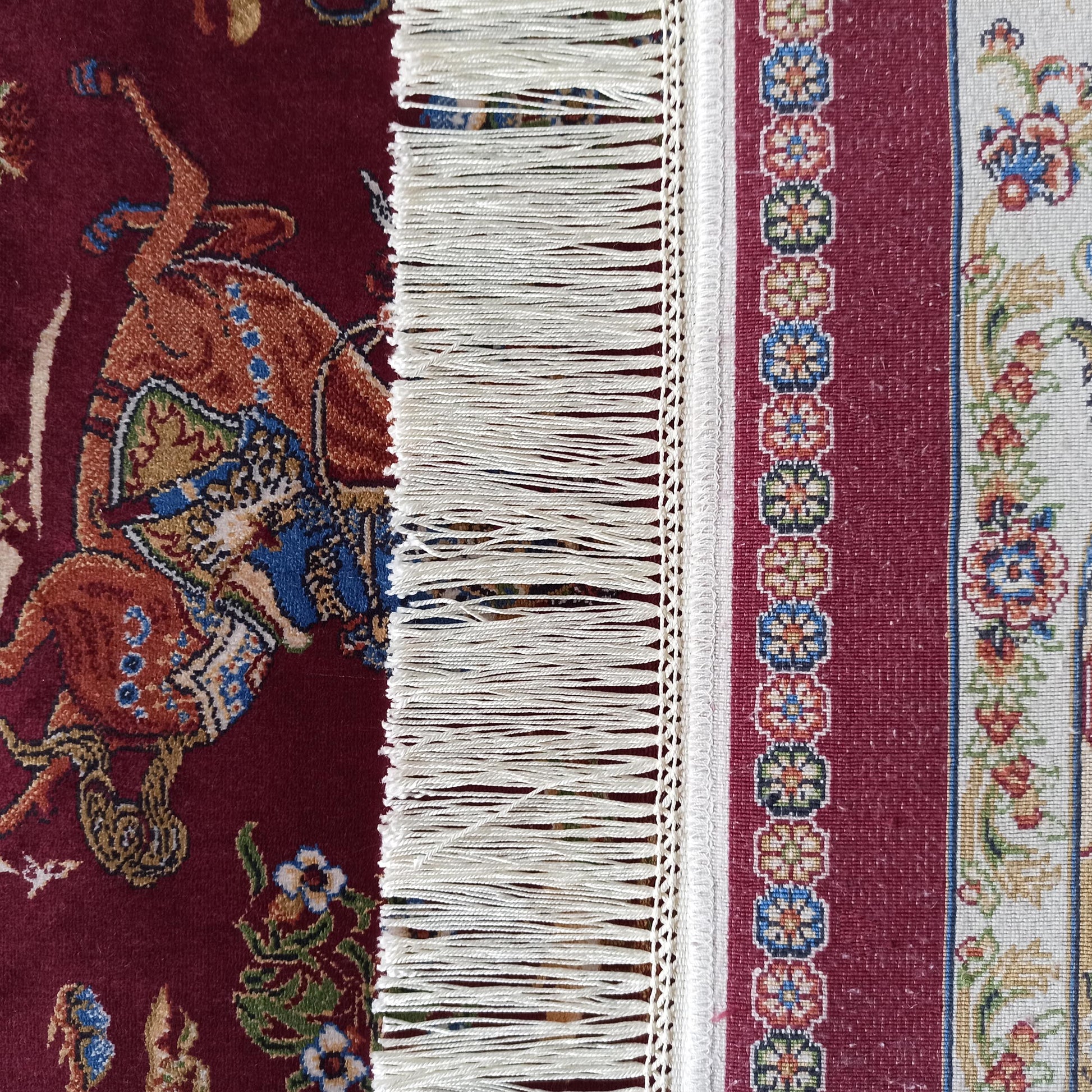 Tapis coloré présentant des motifs orientaux. On distingue des animaux stylisés sur fond bordeaux, des franges blanches et une bande florale crème