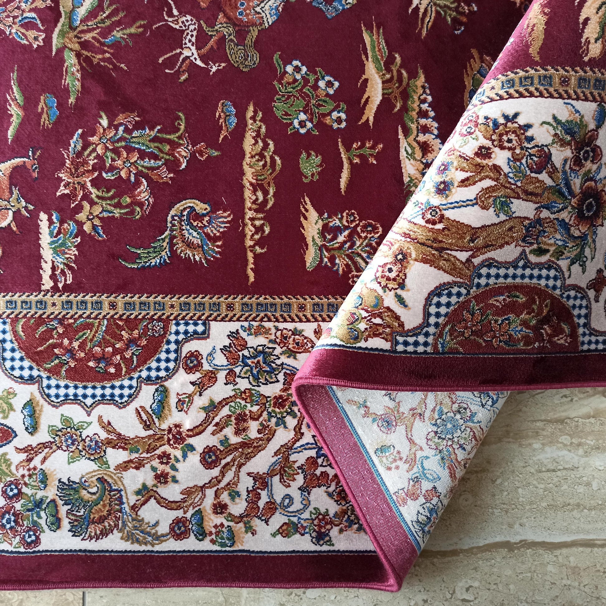 Tapis bordeaux aux motifs floraux et animaliers complexes en crème, or, vert et bleu, partiellement roulé sur un sol clair