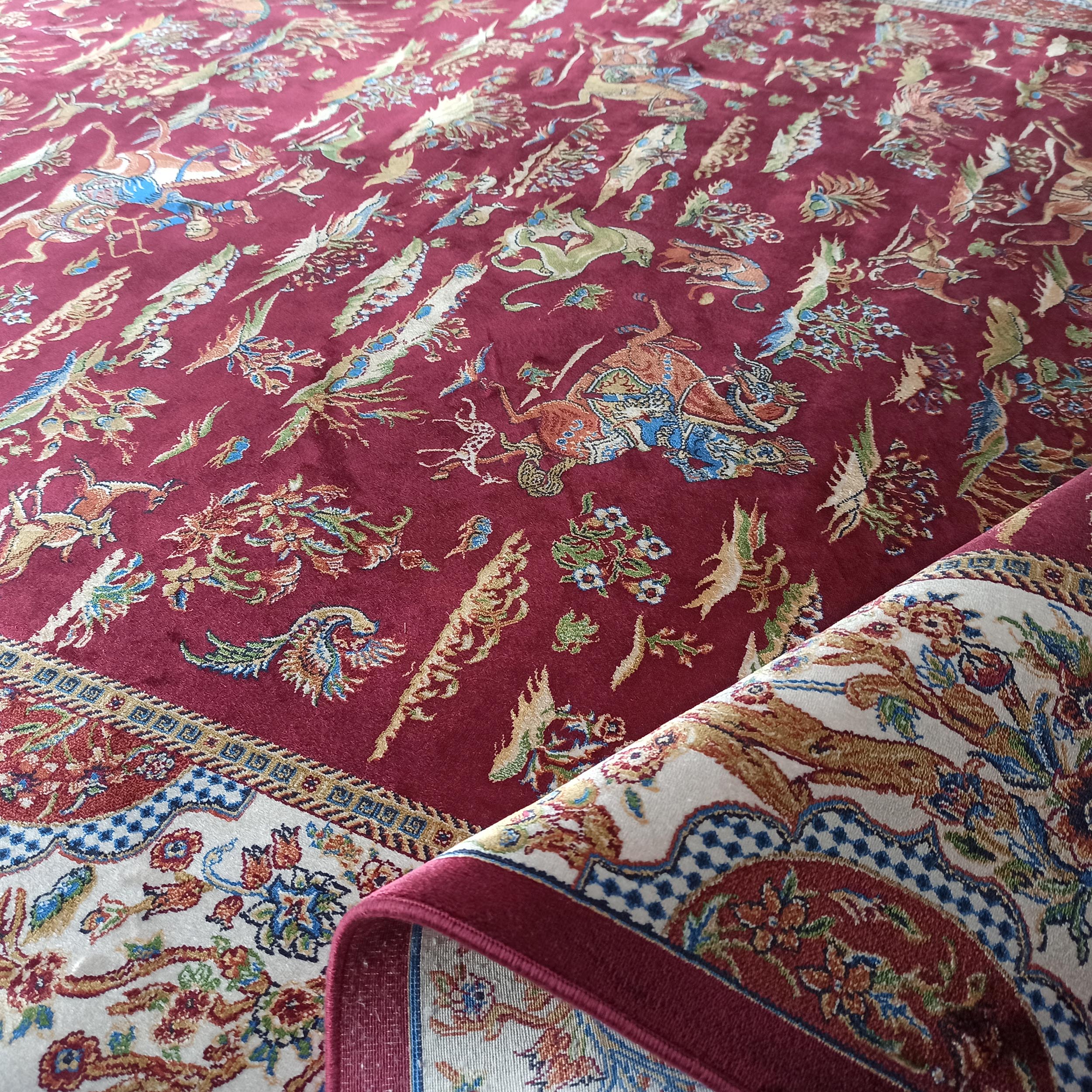 Tapis rouge bordeaux avec un riche motif floral et animalier coloré, bordé d'un motif crème et bleu, partiellement enroulé
