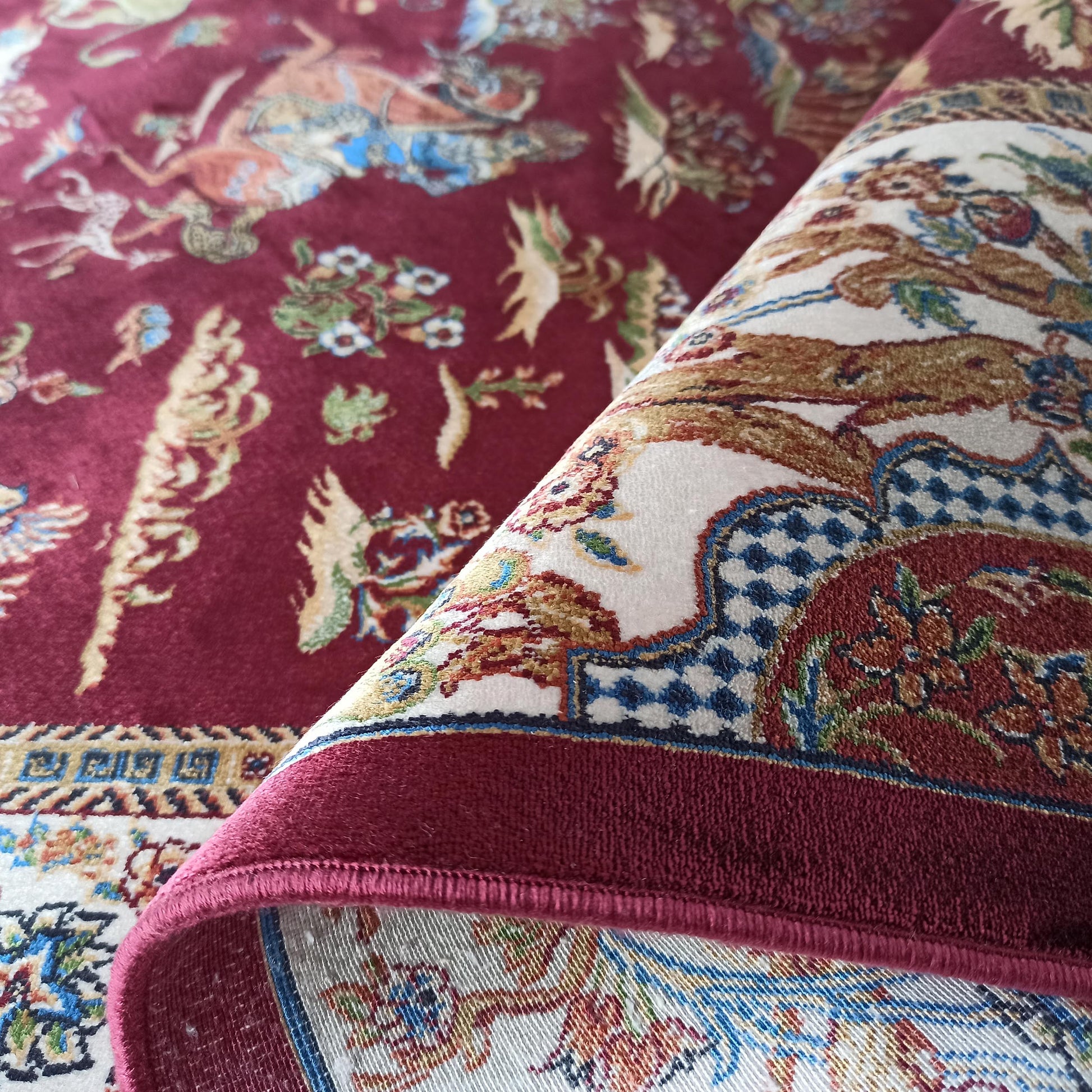 Tapis bordeaux aux motifs orientaux complexes de fleurs et créatures. La section roulée expose une bordure crème, bleue et dorée