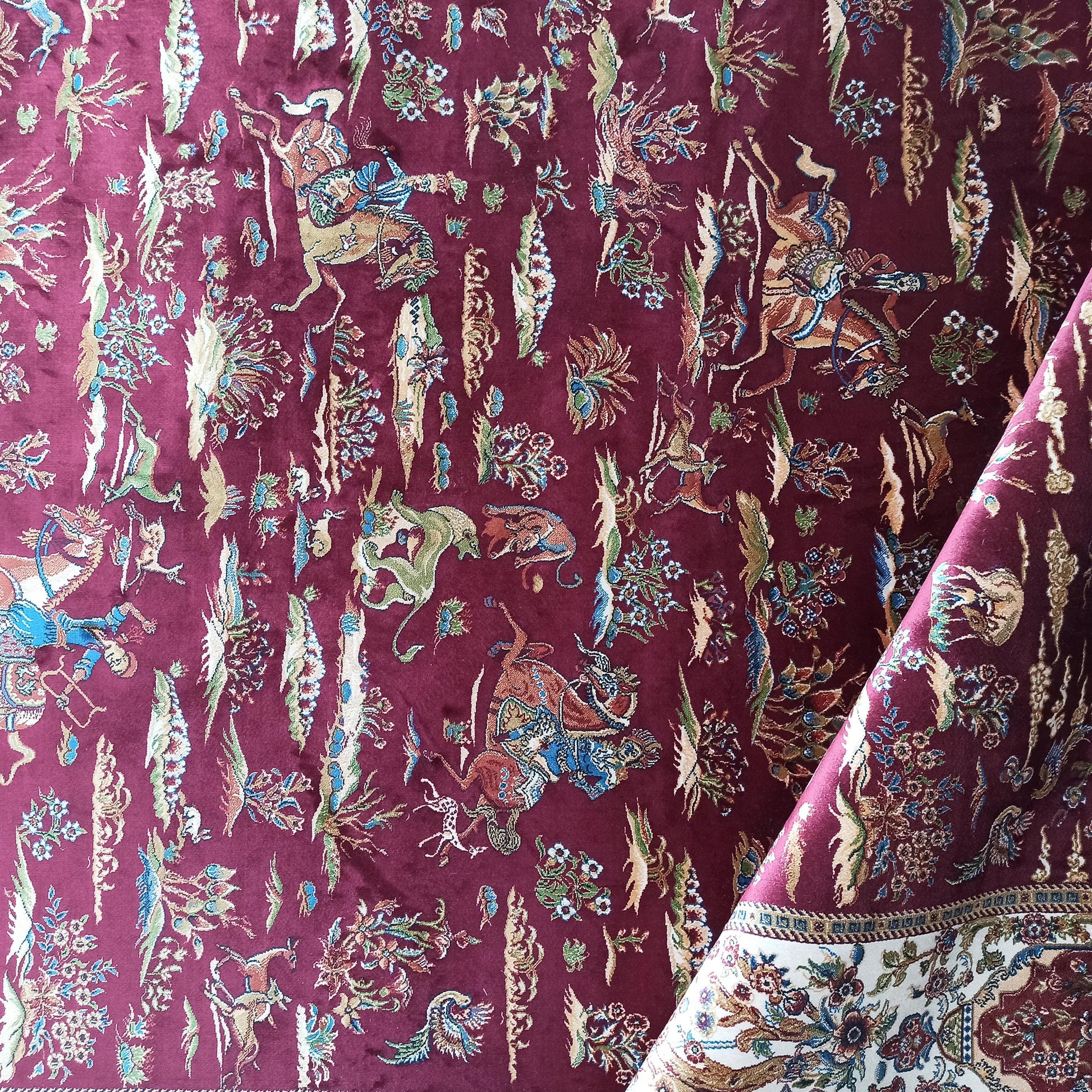 Tapis bordeaux à motifs traditionnels détaillés, figuratifs et floraux. Sa bordure repliée révèle un décor crème et bleu